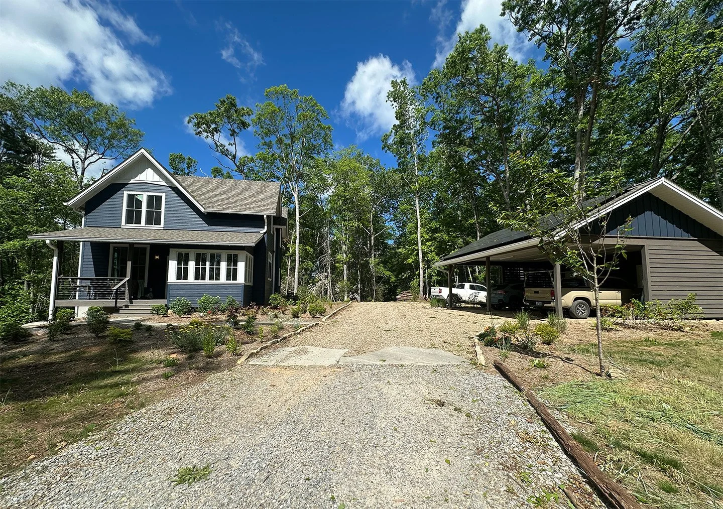 A modern blue two-story house with a front porch, surrounded by small shrubs, greenery, and trees on a clear day with blue skies.