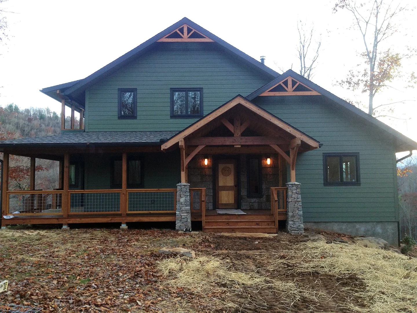 A two-story house with a large front porch, green siding, and black window frames. The house has a wooden gable roof with decorative trim. The front steps lead up to a door with two outdoor lights on either side. The yard is bare with some rocks and 