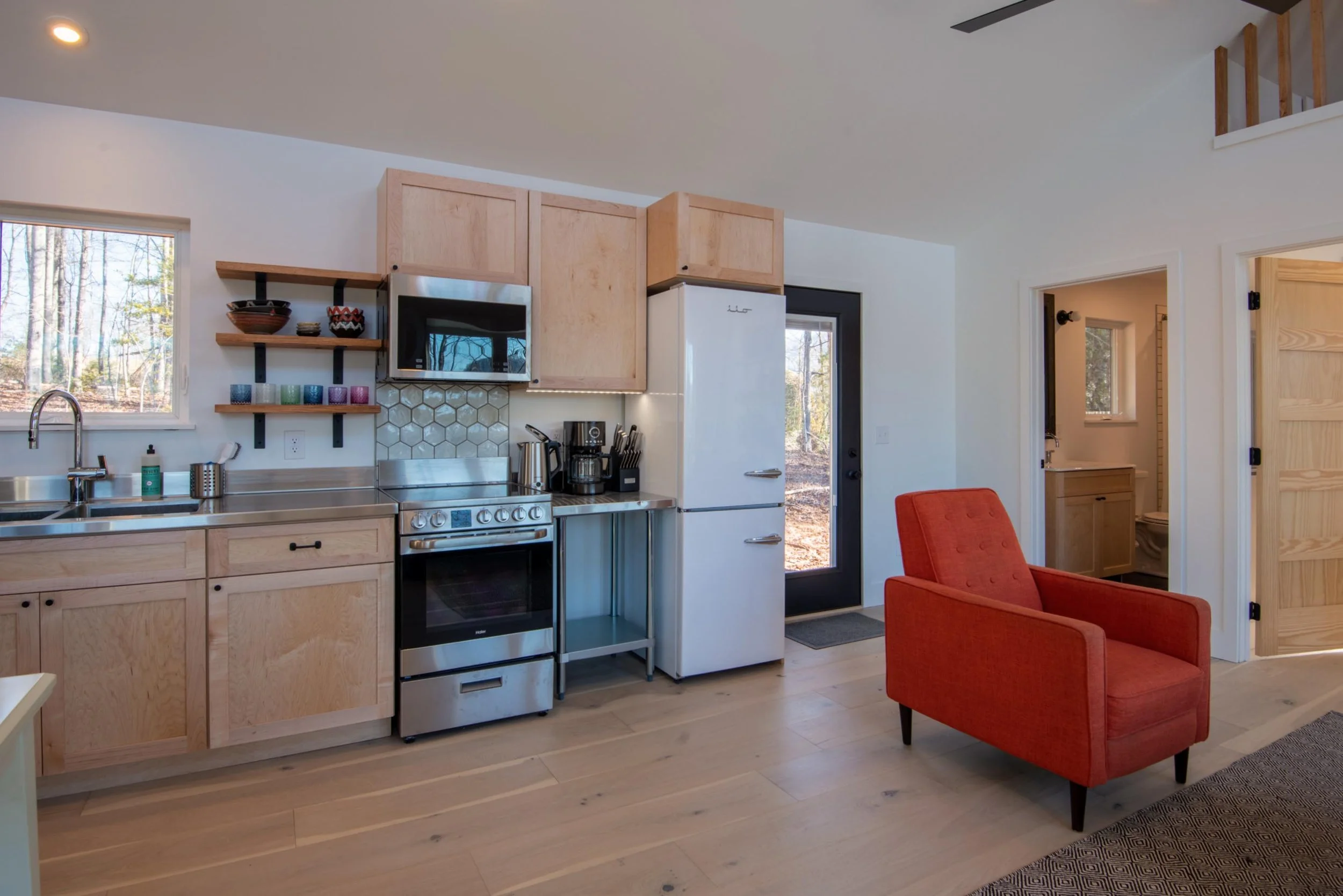 A cozy kitchen and living space with light wood cabinets, stainless steel appliances, and a small orange armchair. A black door with a window leads outdoors, and there is a bathroom visible through an open door.