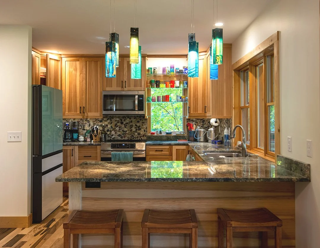 A kitchen with wooden cabinets, colorful hanging pendant lights, a granite countertop, a window with a view of trees, and a breakfast bar with three wooden stools.