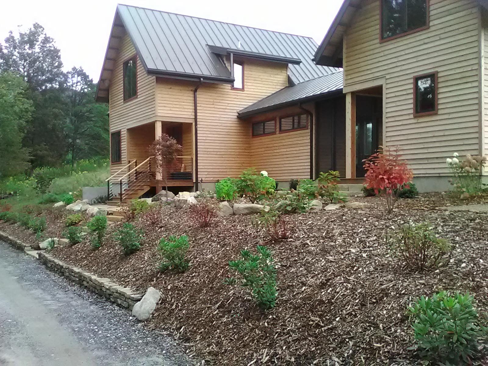 A modern wooden house with a metal roof, surrounded by a landscaped garden with small bushes and flowers.