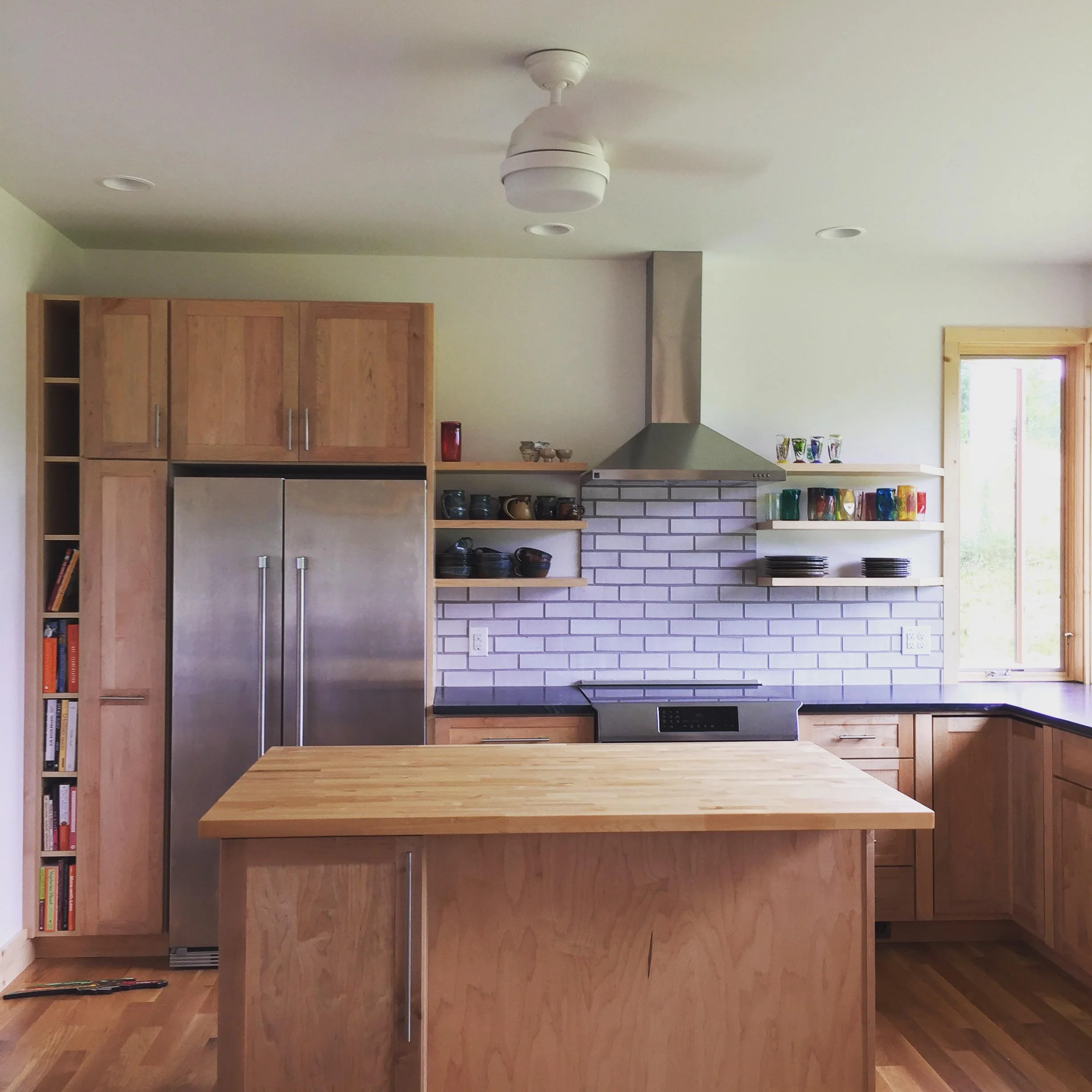 Modern kitchen with light wood cabinets, stainless steel refrigerator, open shelves with bowls and mugs, white brick backsplash, black countertop, and a wooden kitchen island.