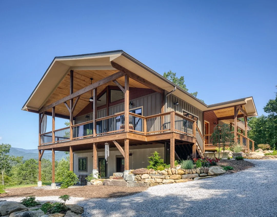 A modern house with a large wooden deck on the upper level, situated on a sloped landscape with rocks and greenery, under a clear blue sky.