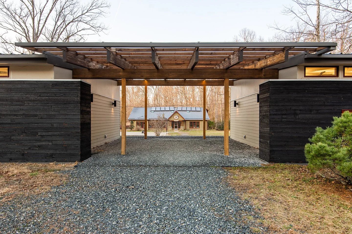 Modern carport with wooden beams and gravel driveway, house with metal roof in background, leafless trees surrounding property.
