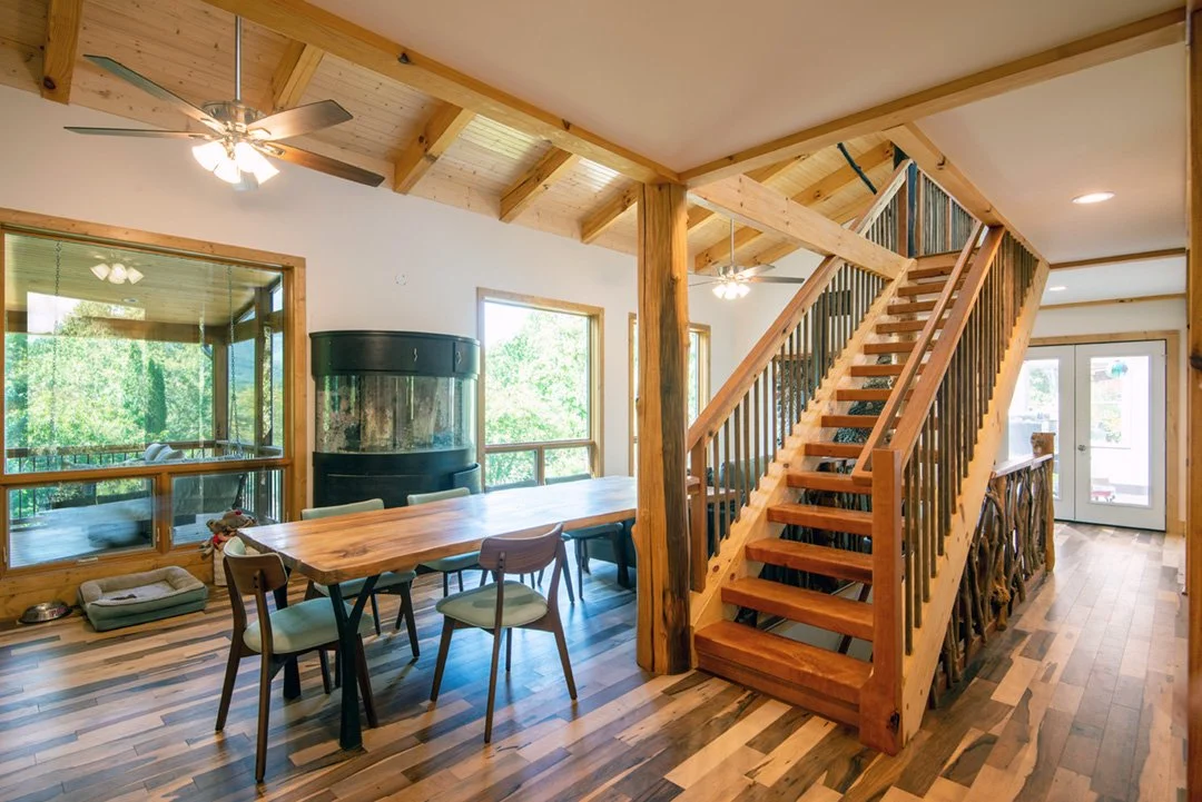 Interior view of a wooden cabin with a staircase, dining area, wood flooring, and large windows with a view of greenery outside.