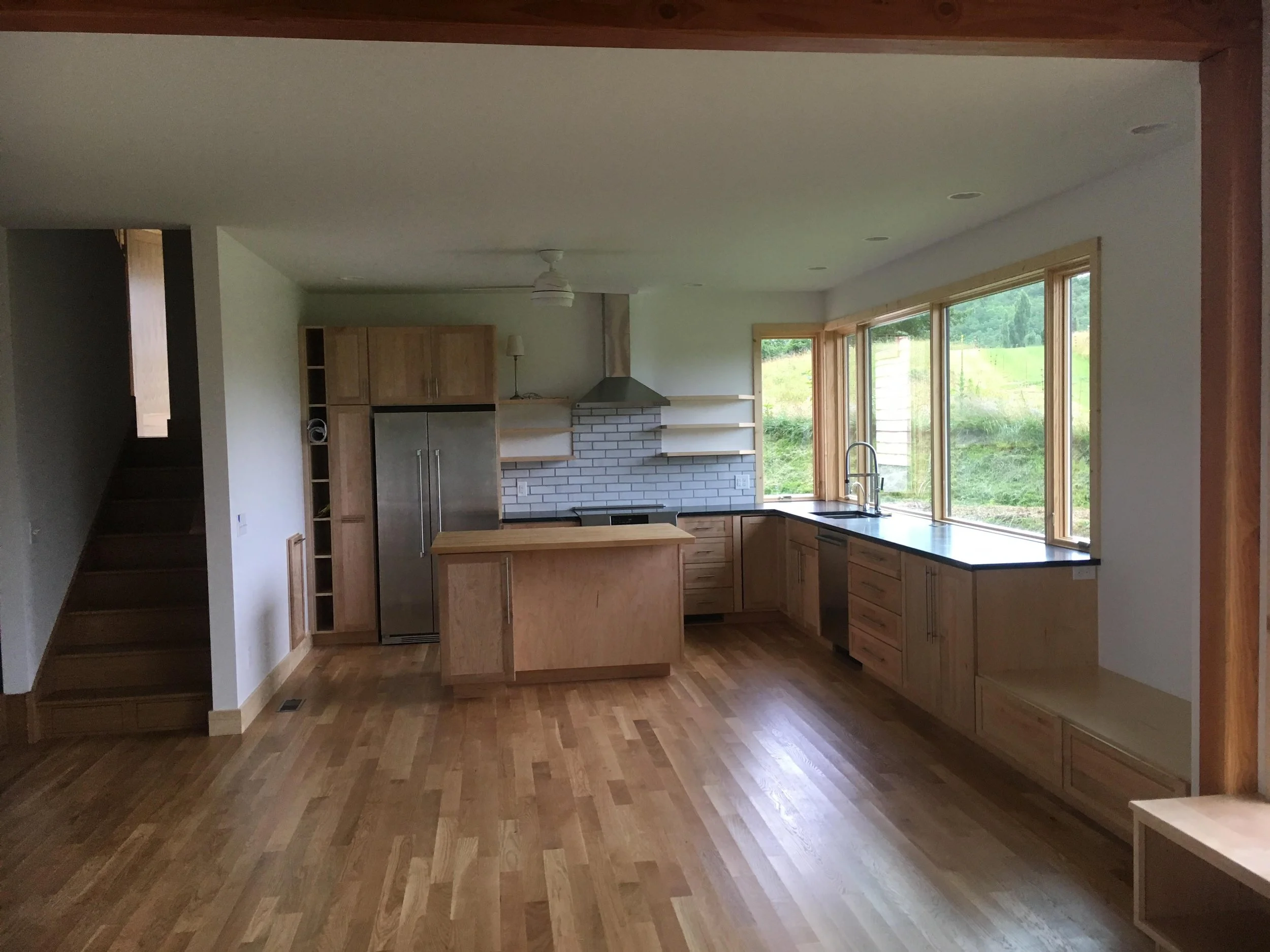 Unfinished kitchen with natural wooden cabinets, large windows, hardwood flooring, white subway tile backsplash, and a modern stainless steel refrigerator.