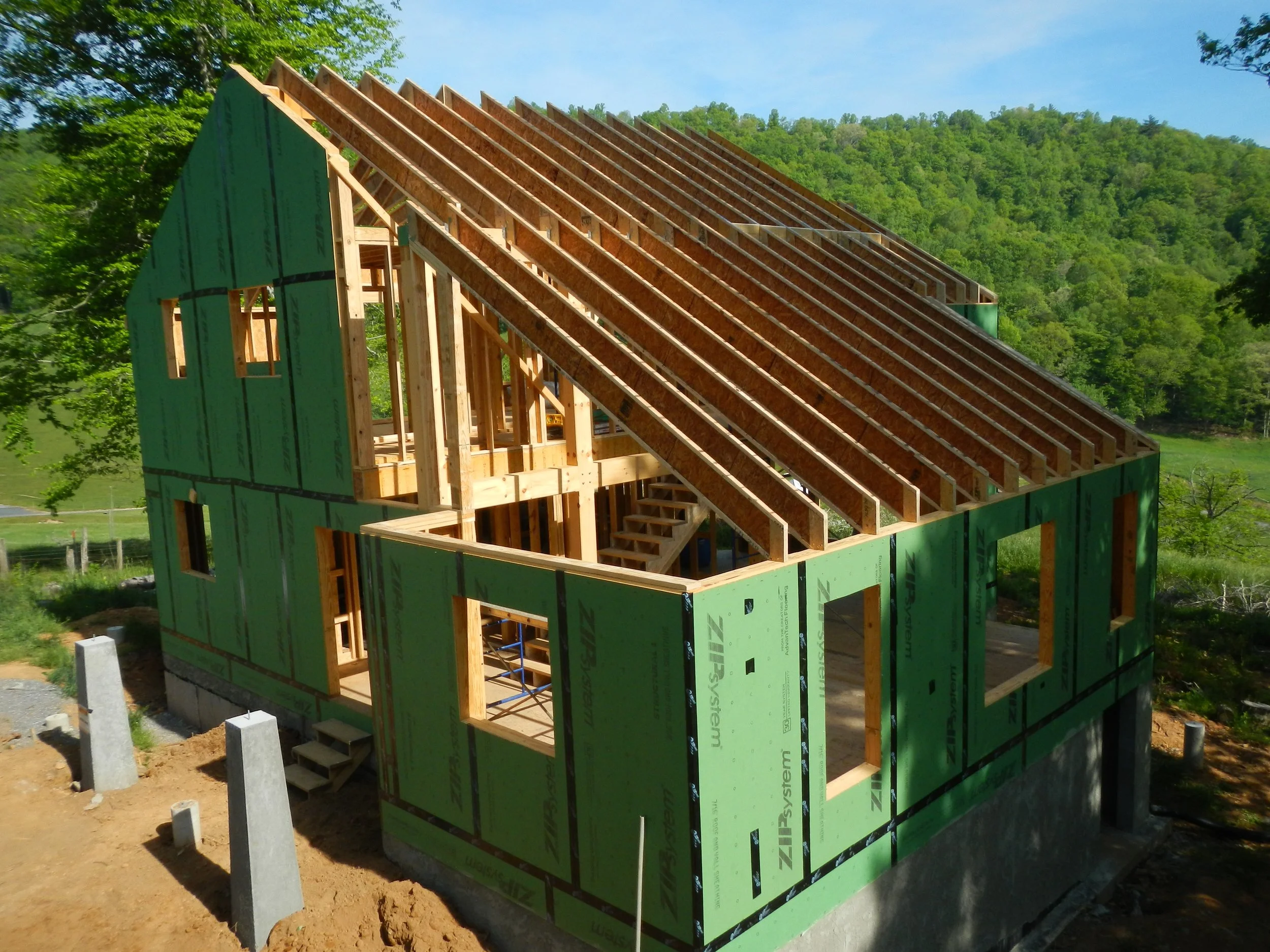A house under construction with wooden framing and green sheathing, set in a green landscape with trees and a hill in the background.