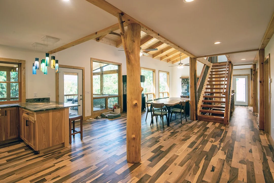 Open-concept living room and kitchen with wooden accents, large windows, a dining table, staircase, and hardwood floors.