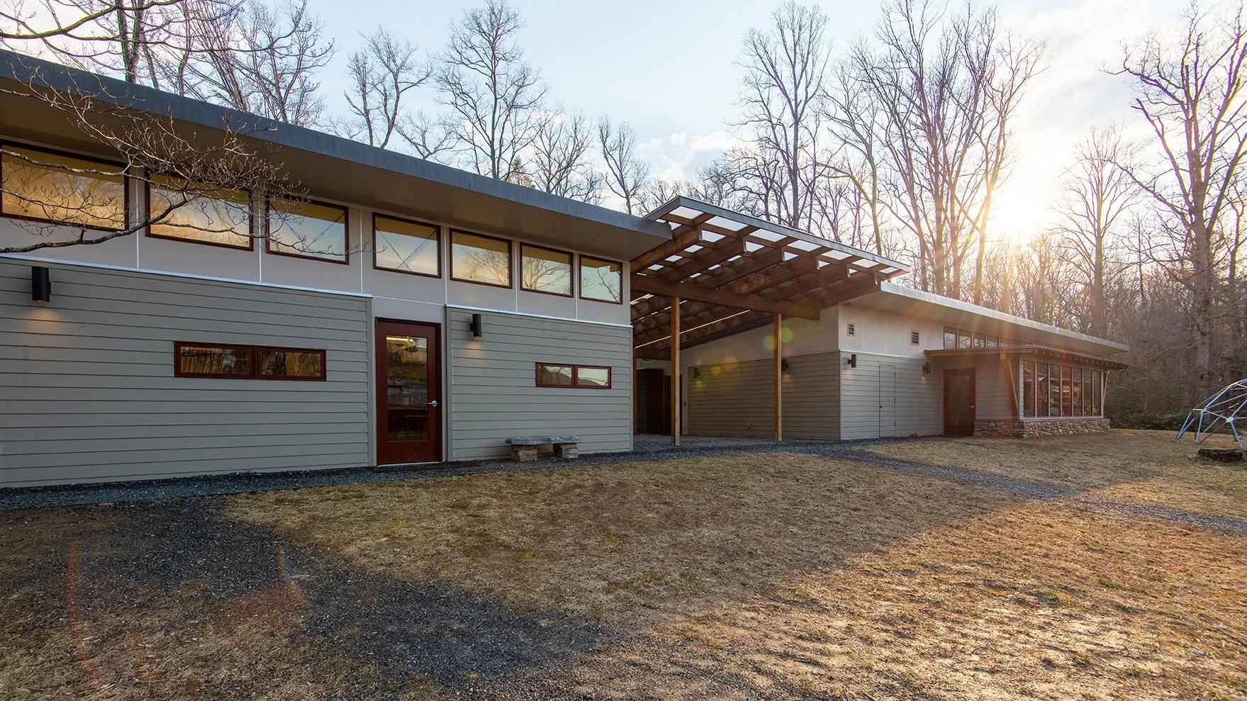 Modern building with horizontal siding, multiple windows, and a covered patio, surrounded by leafless trees and a grassy yard during sunset.