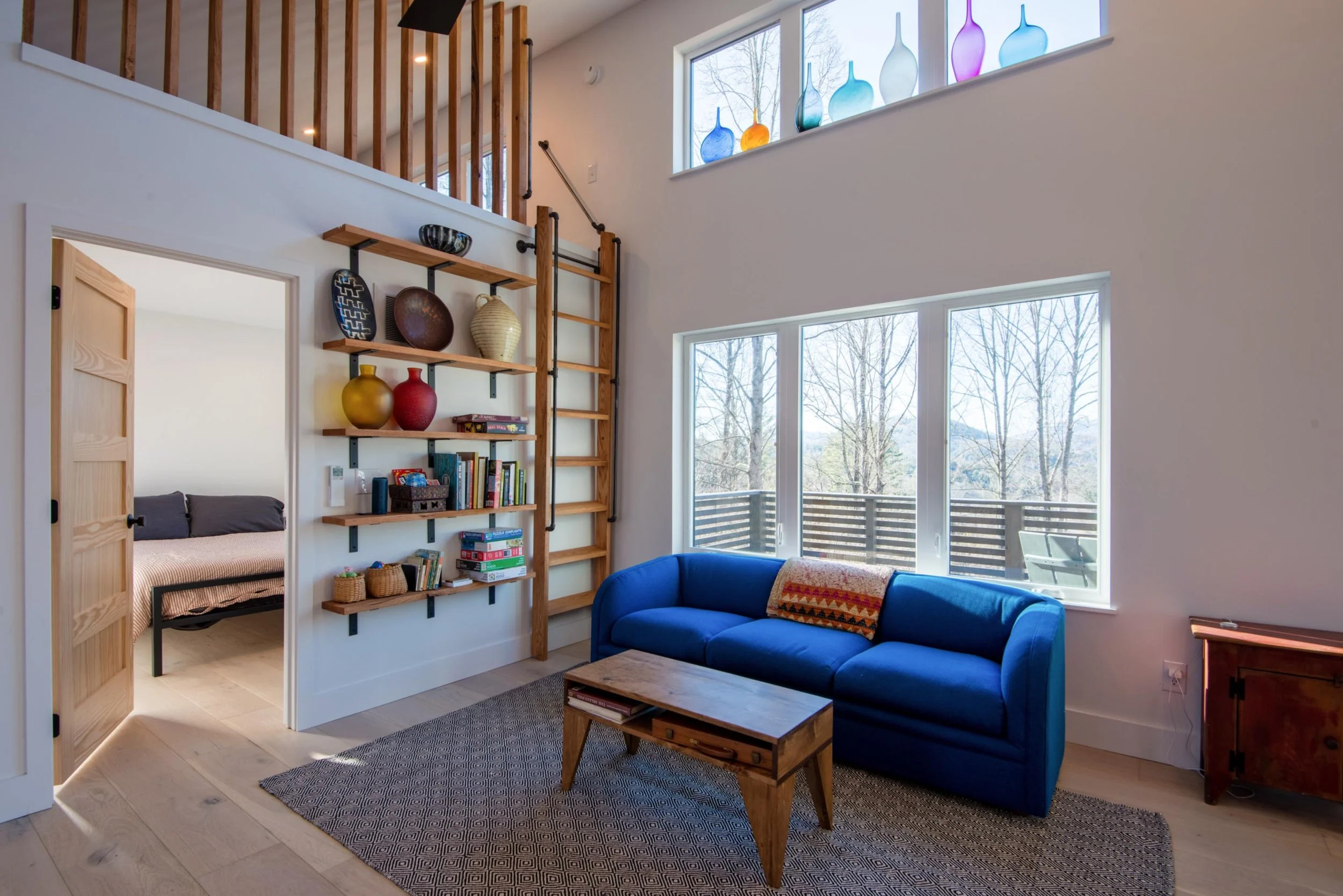 Living room with large windows, blue sofa, wooden coffee table, and a wall-mounted wooden ladder with shelves holding decorative vases, books, and baskets. Partial view of a bedroom through an open door.