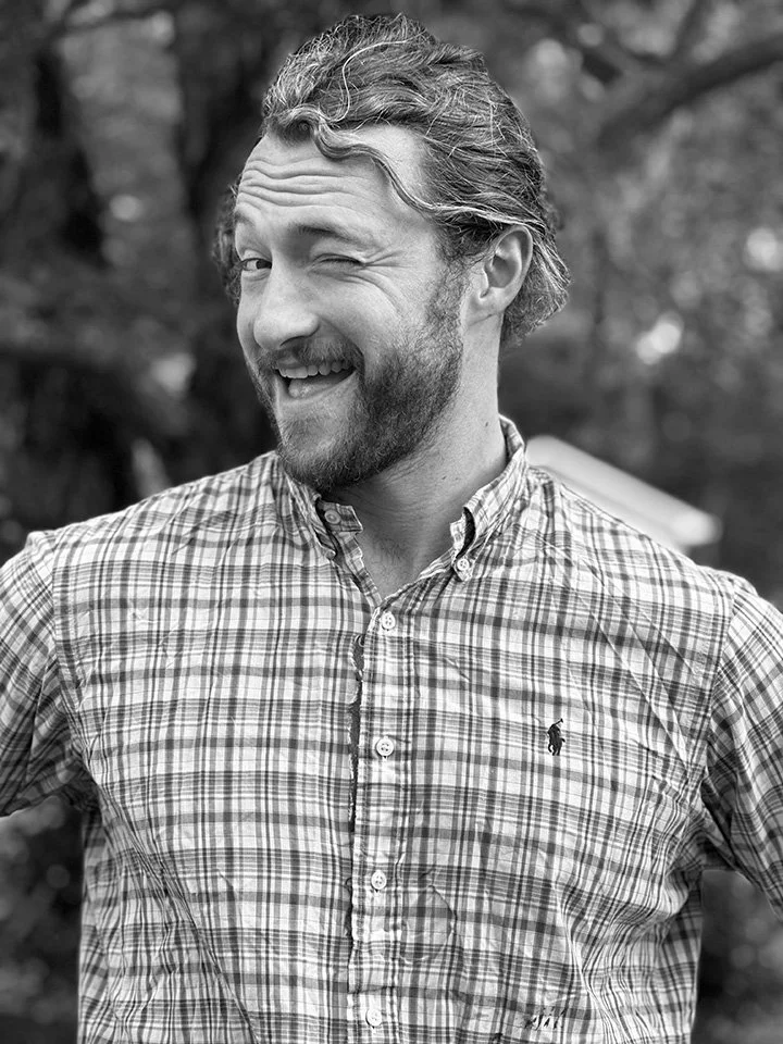 A man with a beard and wavy hair, smiling and winking, wearing a plaid button-up shirt outdoors.
