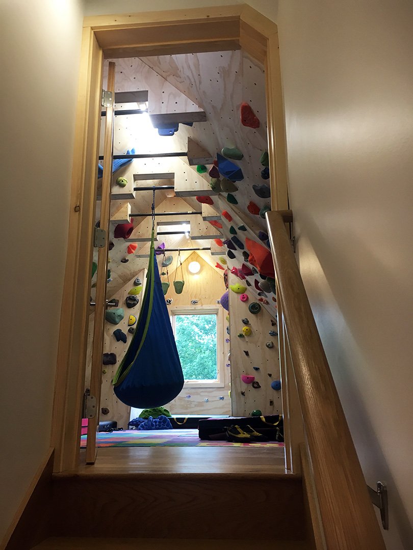 Indoor climbing wall with various colored holds, a blue hanging sling, and a window showing trees outside.