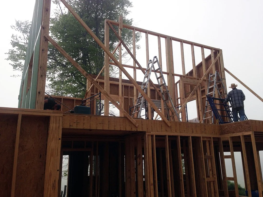Construction workers building the wooden frame of a house, with ladders set up and trees in the background.
