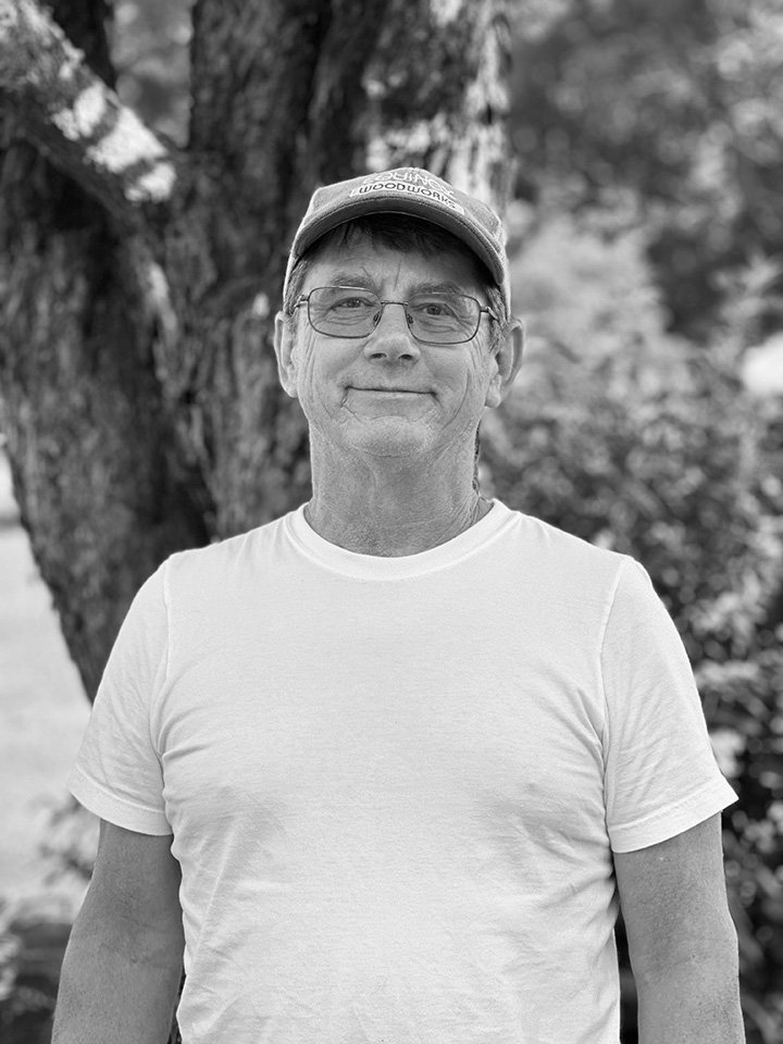 A middle-aged man wearing glasses, a cap, and a white t-shirt is smiling outdoors in front of a tree.
