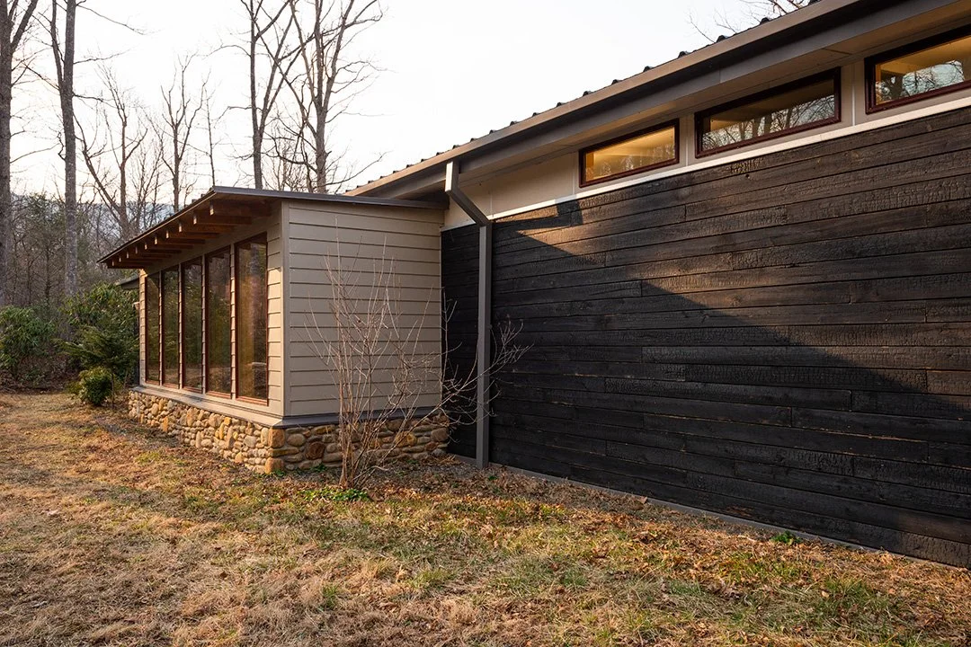 Side view of a modern house with beige siding, a stone foundation, and black horizontal wood paneling. The house has large windows and is surrounded by leafless trees and a grassy yard.