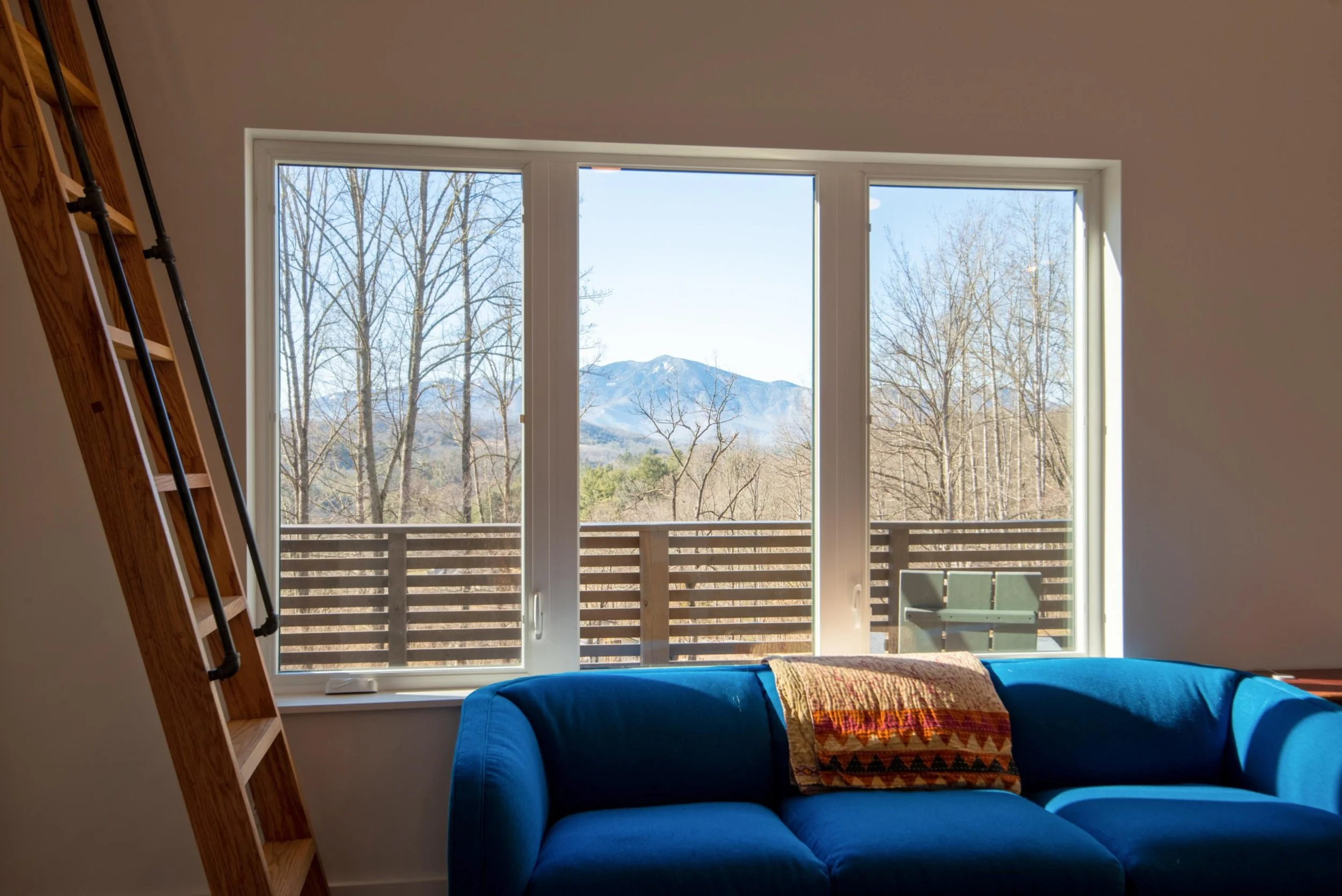 Living room with large window showing mountain view, blue sofa with blanket, wooden ladder to the left, and a balcony with a green chair outside.