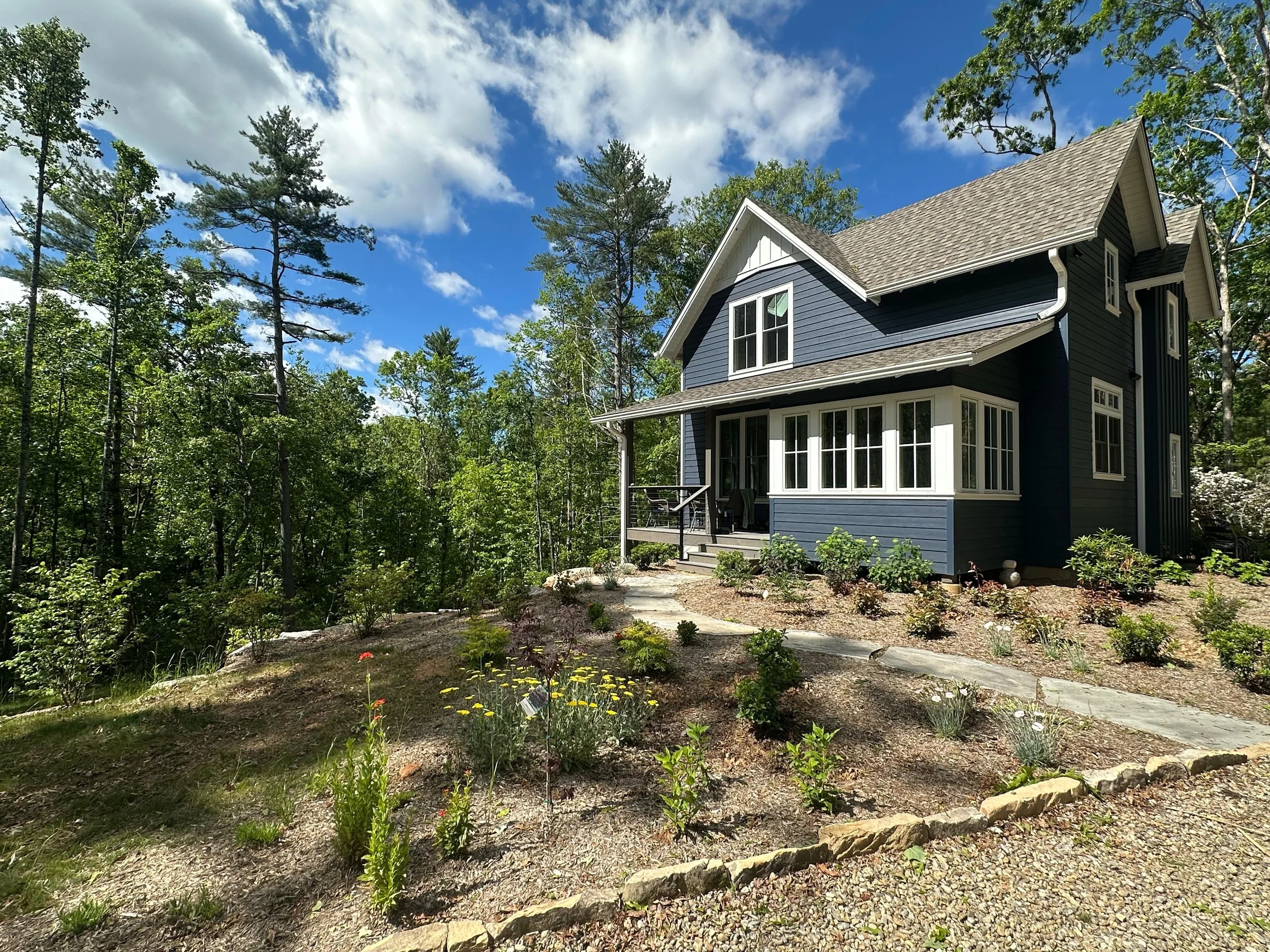 A blue, two-story house with white trim and a front porch, surrounded by trees and a landscaped yard with small plants and a stone pathway under a partly cloudy sky.