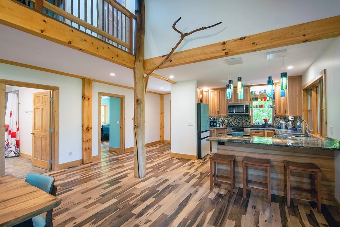 Open-concept kitchen and living area with wooden beams, a tree in the center, and hardwood floors. Kitchen features wood cabinets, a window with colorful hanging pendant lights, and a granite island with three stools.