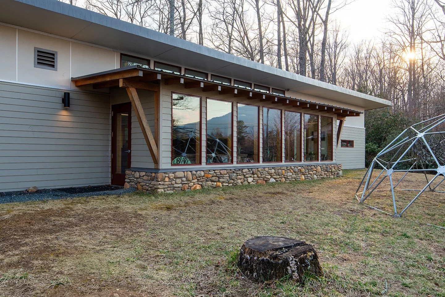 Modern house with large glass windows, stone foundation, and wooden support beams, set against a backdrop of trees and a setting sun.