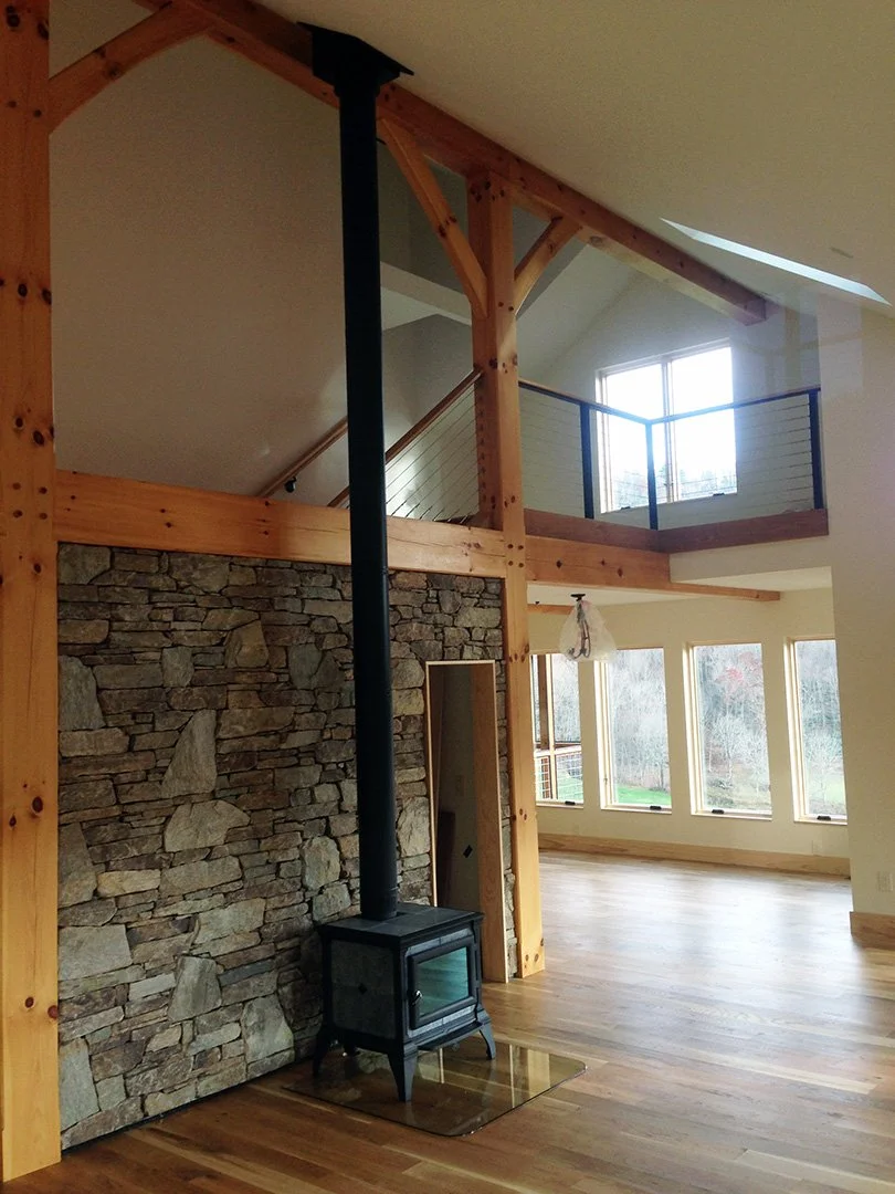 Interior of a house with wood floors, a stone accent wall, a black wood stove, and a loft area with a railing and large windows.