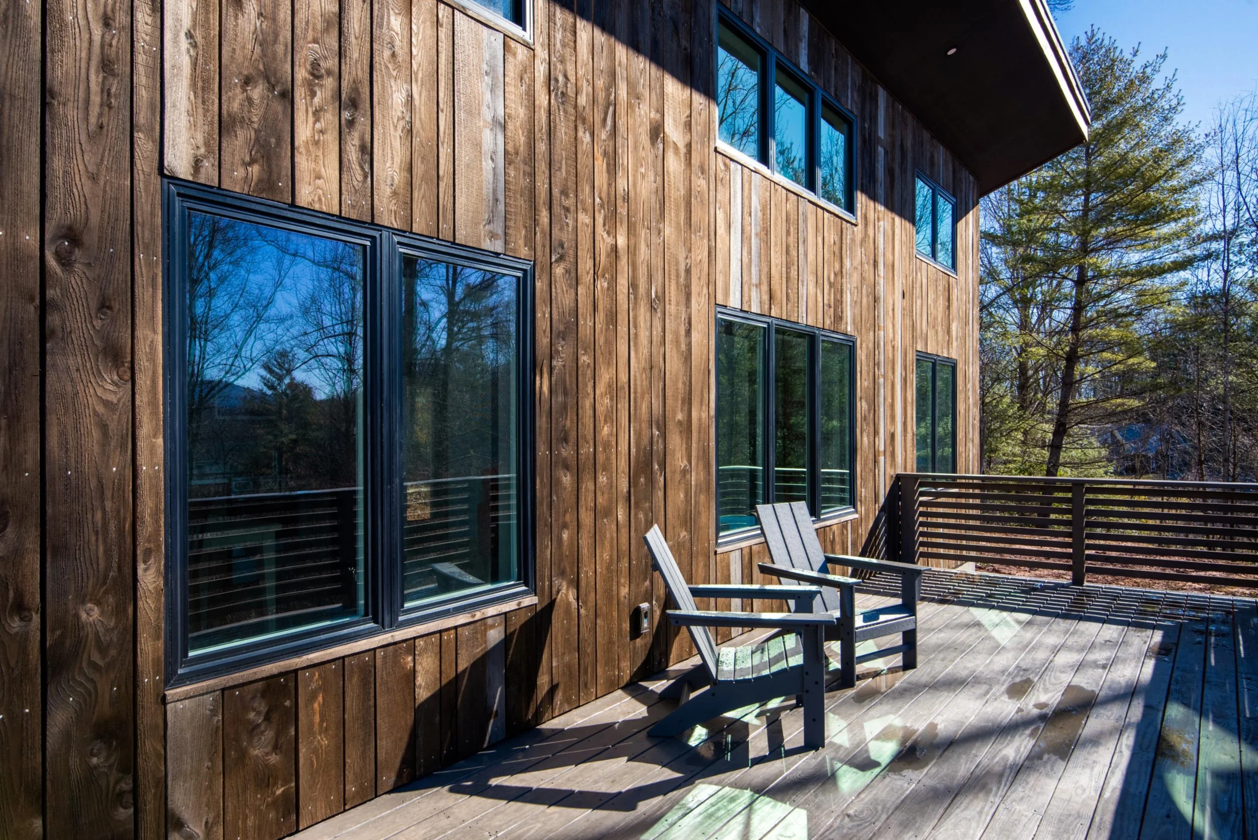 A wooden deck with two Adirondack chairs and a railing, attached to a building with large windows and a wooden exterior, surrounded by trees.