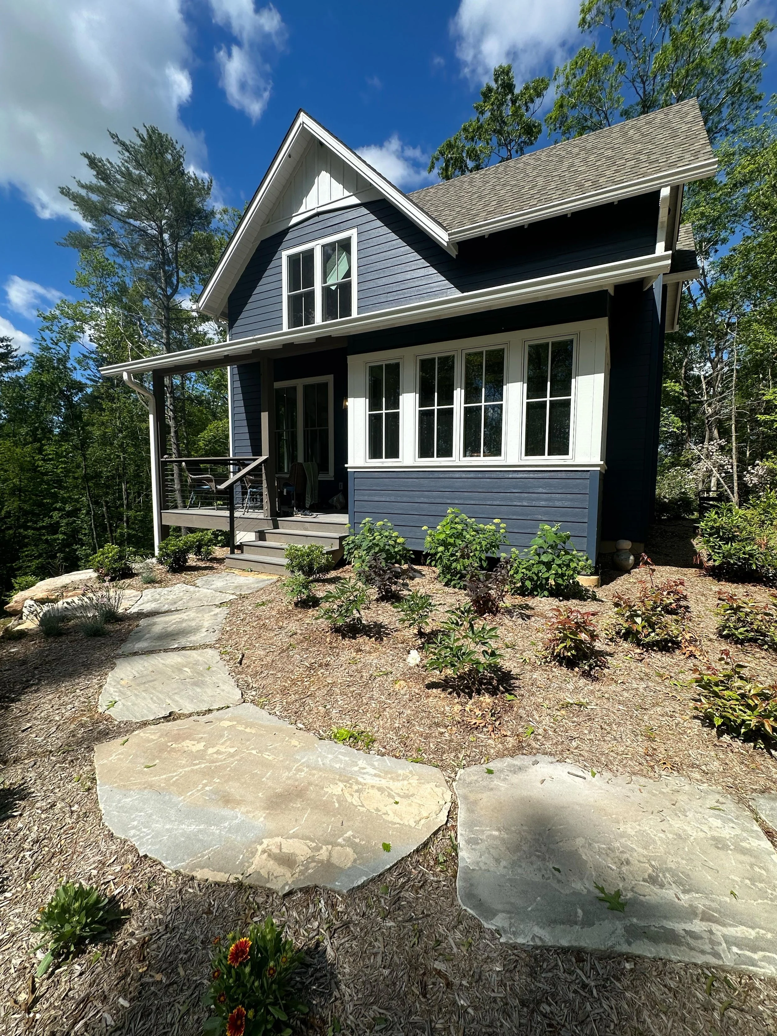 A two-story house with dark blue siding, white framing, and a gray shingle roof, surrounded by trees, with a stone pathway leading to the front porch that has stairs and a railing, and landscaped plants in the yard.