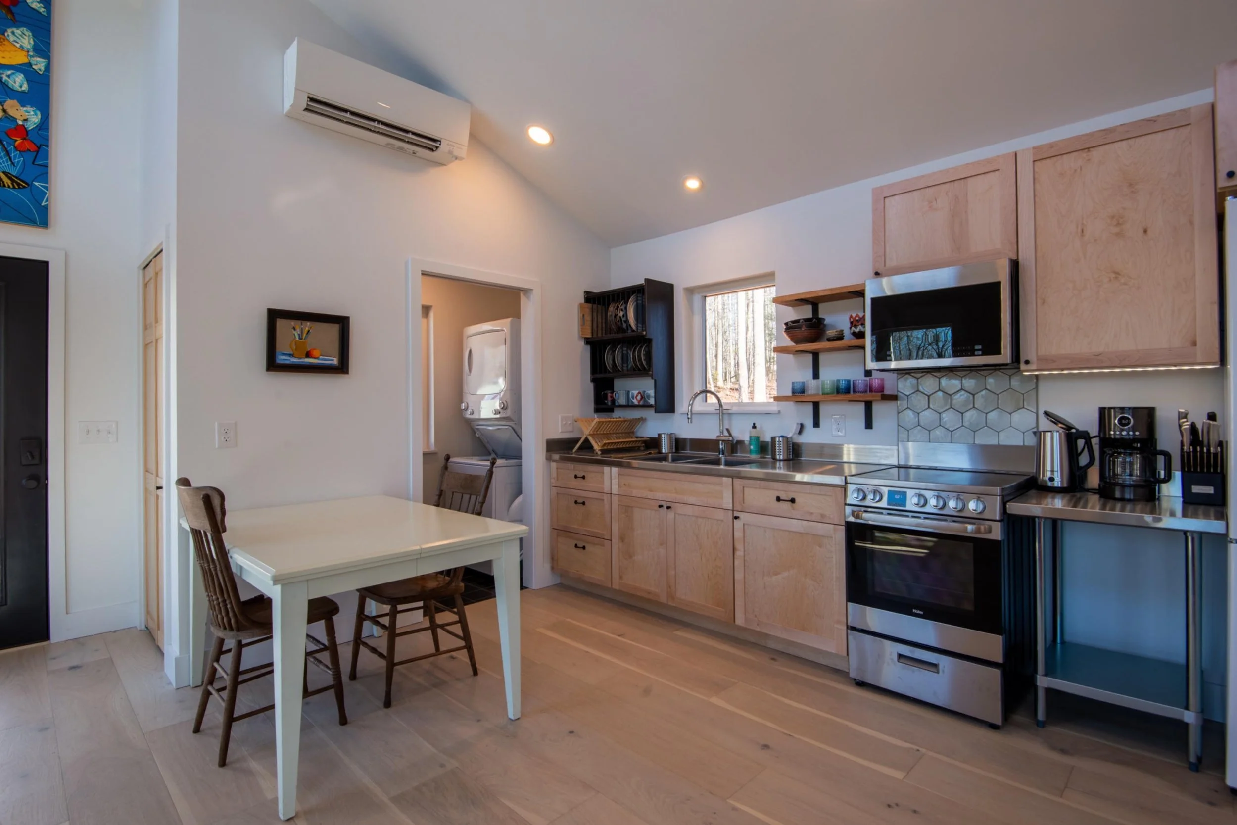 Modern kitchen with light wood cabinets, stainless steel appliances, and open shelving with colorful cups. Small dining table with two chairs, laundry area with stacked washer and dryer, and a window showing trees outside.