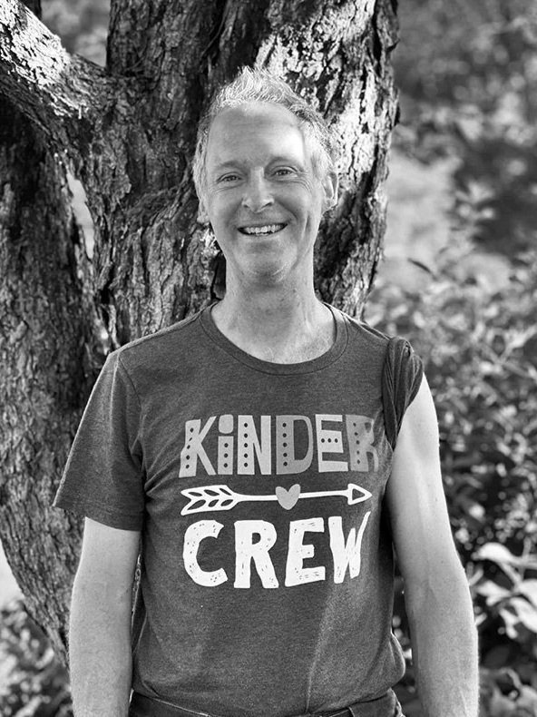 Smiling woman standing outdoors in front of a tree, wearing a T-shirt that says "Kindness Crew" with an arrow and heart design.