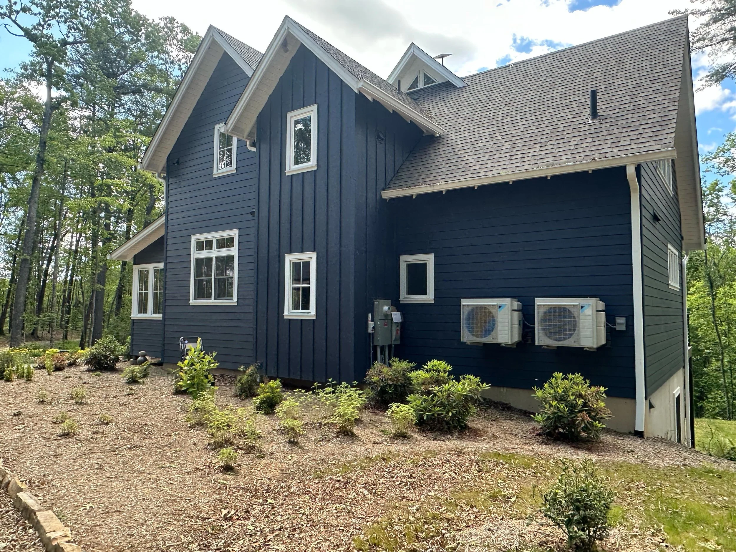 Blue house with multiple windows and air conditioning units on the side, surrounded by trees and a garden with small bushes and gravel ground.