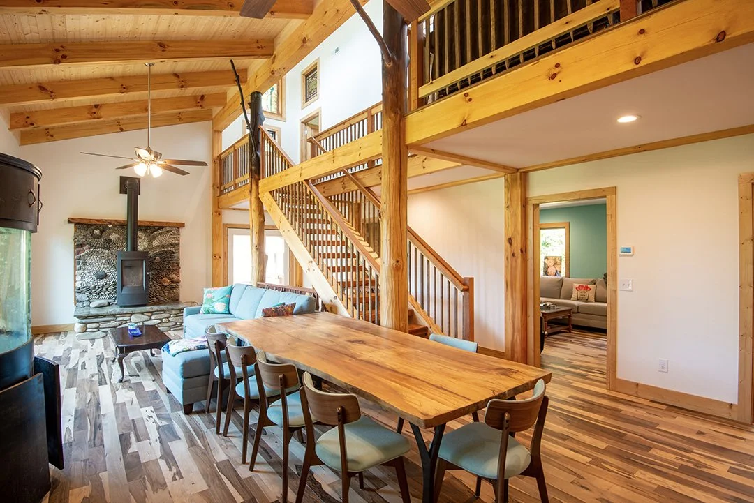 Interior of a wooden house with a high ceiling, wooden beams, a staircase, and a loft overlooking the living space. There is a dining table with chairs, a sofa with cushions, a floor lamp, and a stone fireplace with a wood stove.