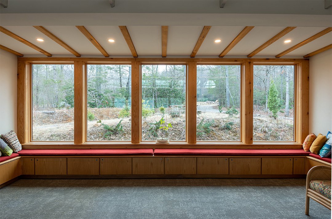 A large window with wooden framing overlooking a wooded yard, featuring built-in seating with red cushions and colorful pillows, adjacent to a small chair with patterned upholstery.