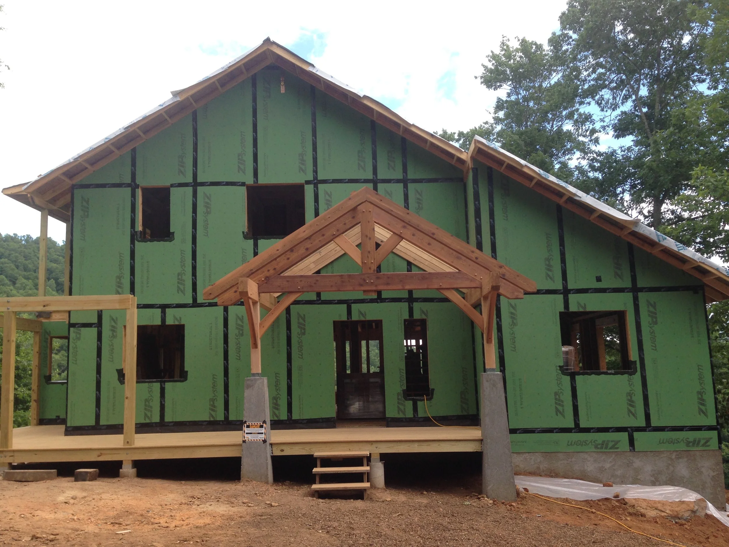 A house under construction with green sheathing, a wooden front porch with a gabled roof, and multiple windows. The house is elevated with concrete foundation pillars and is surrounded by trees.