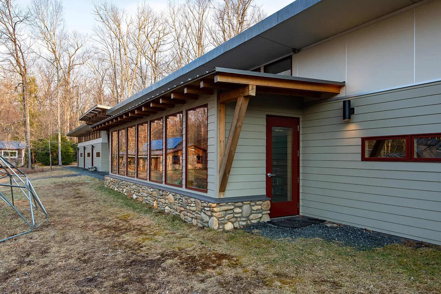 Modern house with large windows, stone foundation, gray siding, and red door, surrounded by trees.