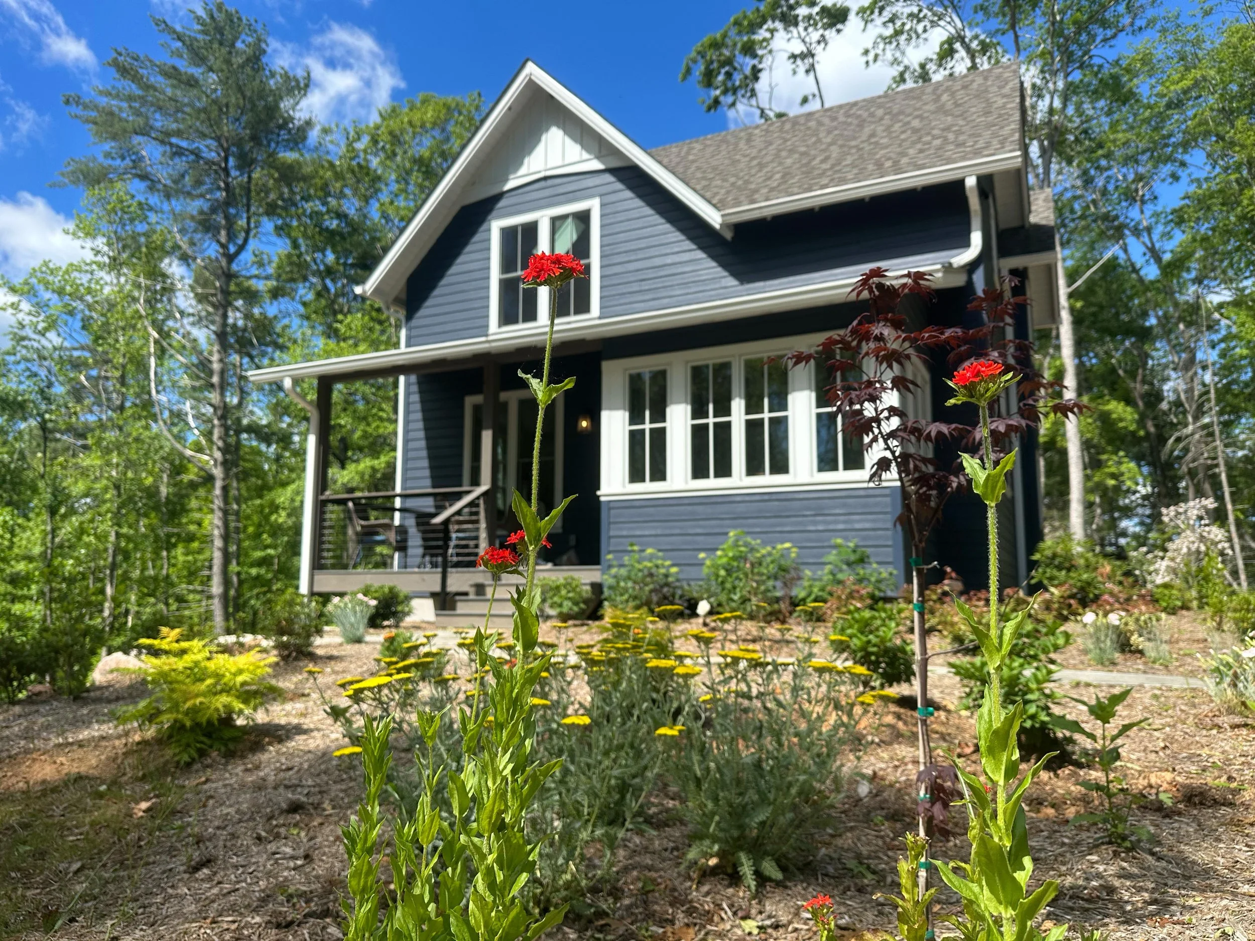 A two-story blue house with white trim and a front porch, surrounded by a garden with red flowers, green shrubs, and trees under a partly cloudy blue sky.