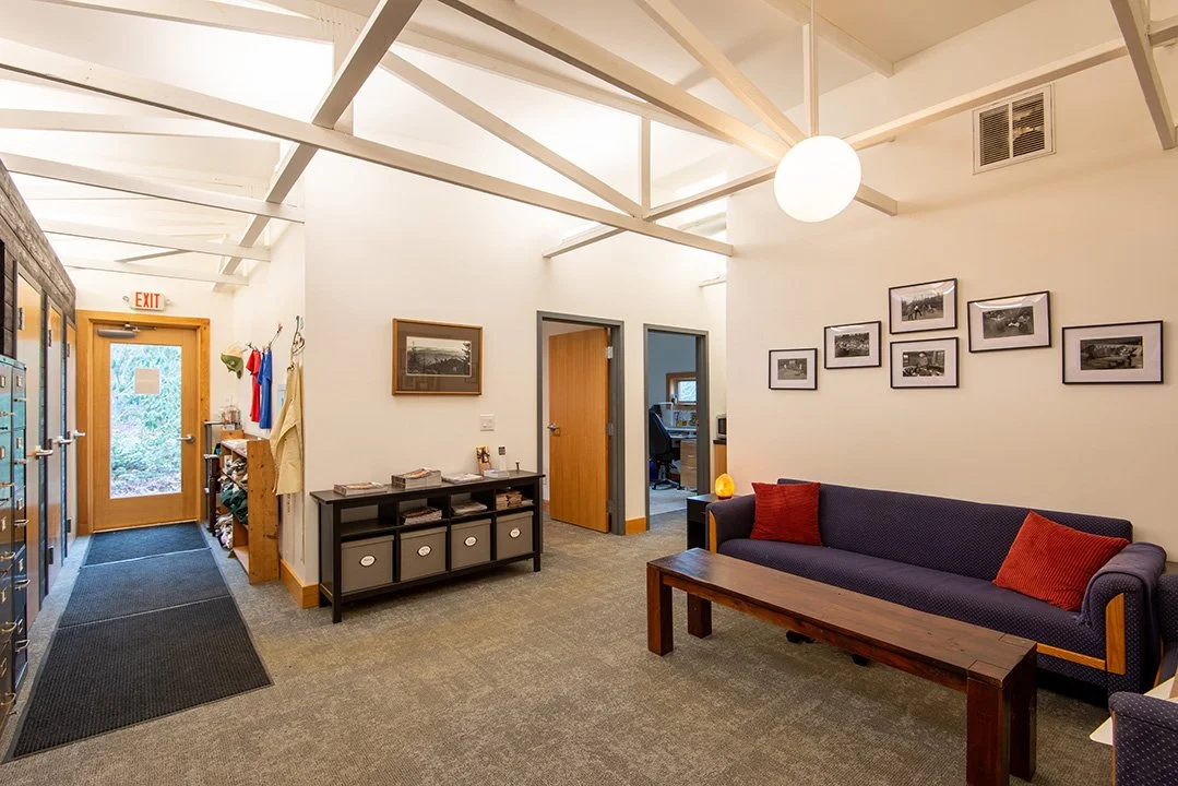 Interior of a waiting room or lobby with a gray carpet, a navy sofa with red pillows, a wooden coffee table, a black console table with storage bins, framed black-and-white photographs on the wall, a round ceiling light, an exit door with a glass win