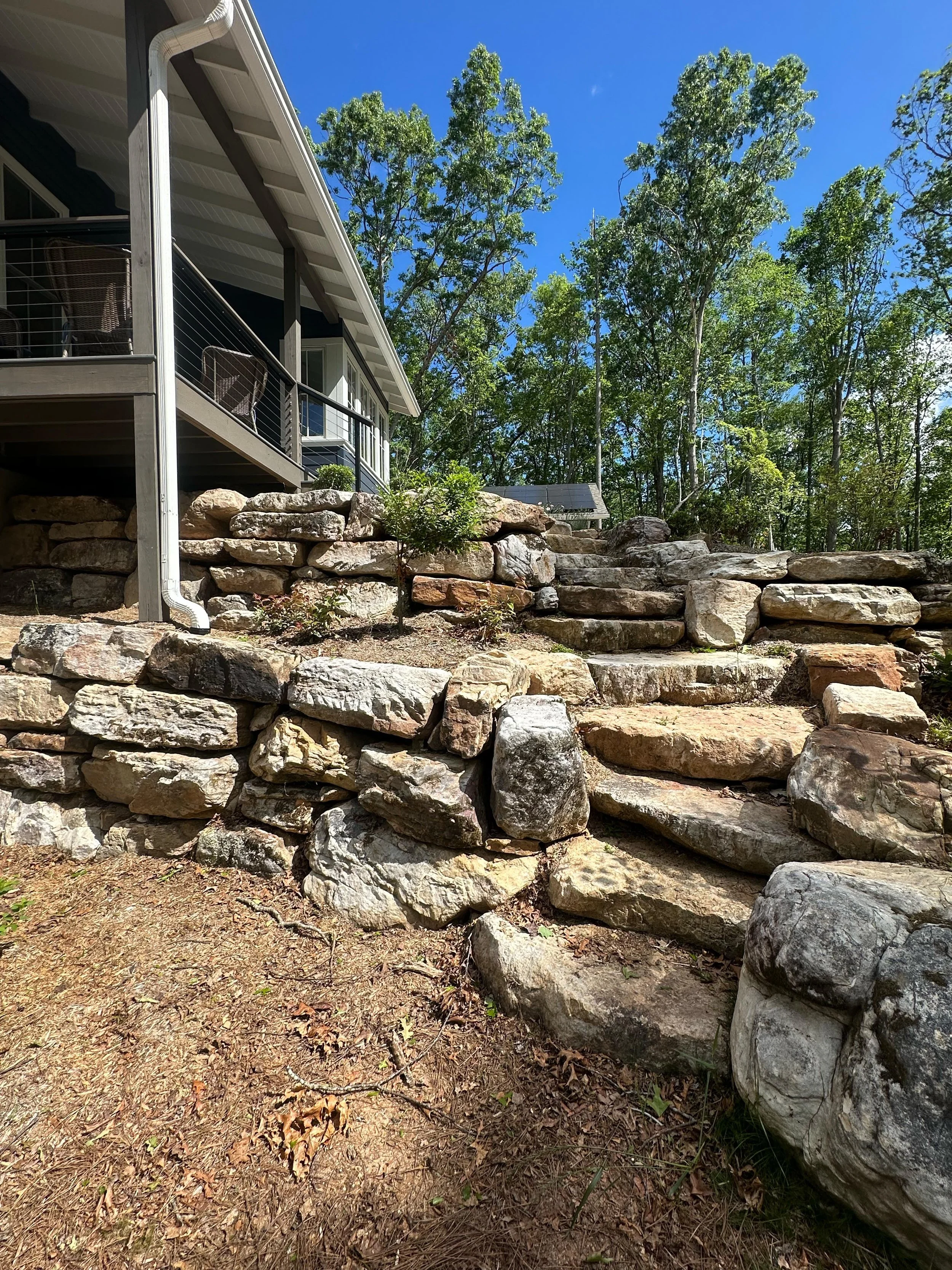 Stone steps leading up a hillside yard with large rocks and a house with a deck in the background, trees surrounding the area, under a clear blue sky.
