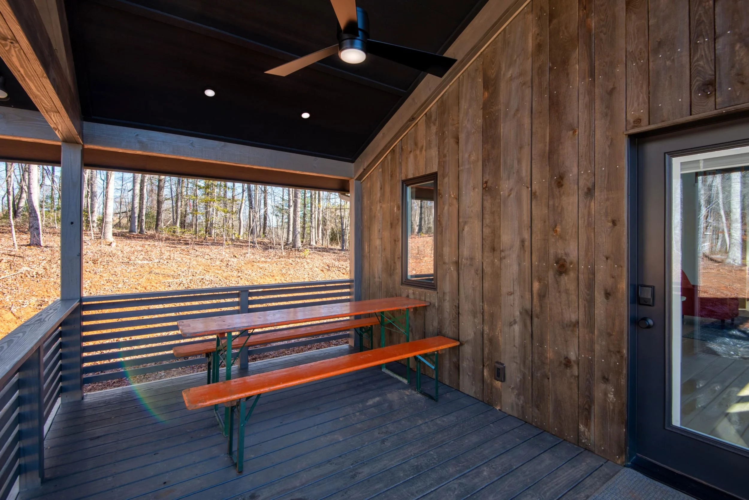 Covered outdoor porch with wooden walls, ceiling fan, and picnic table overlooking a wooded area.
