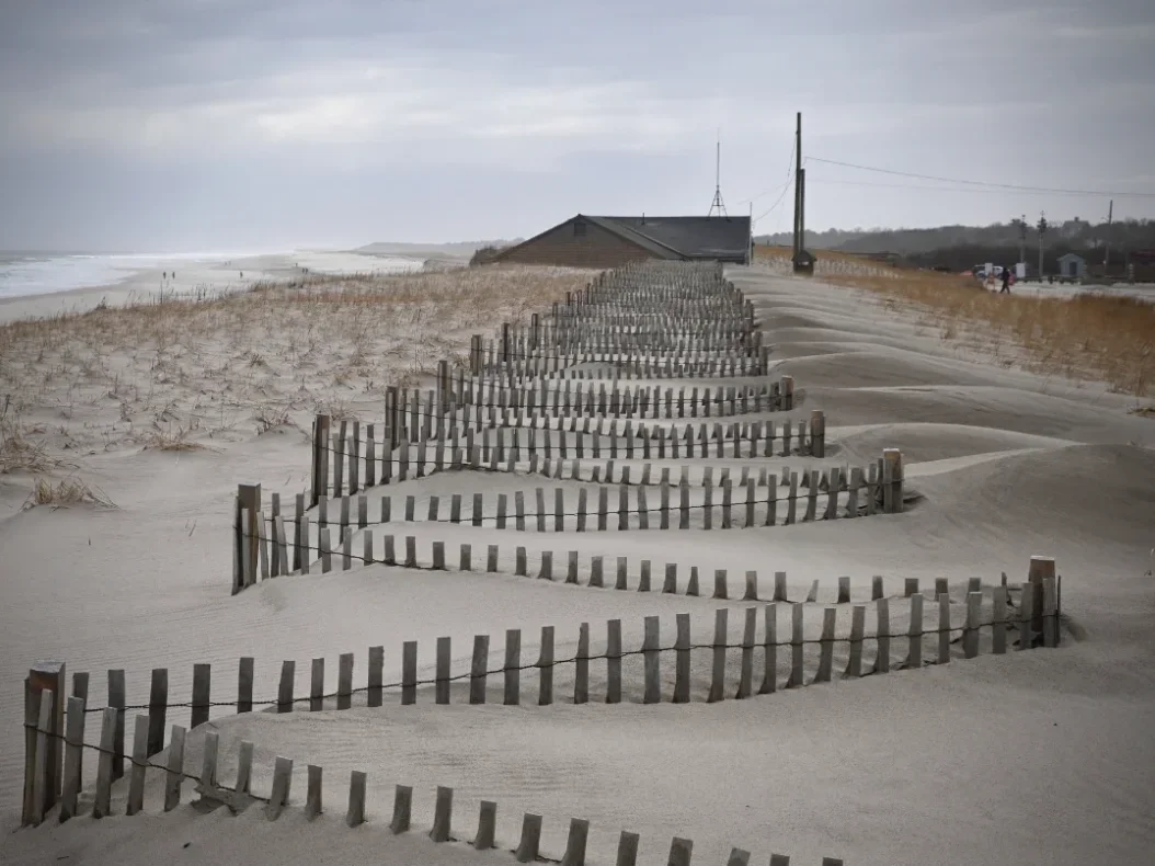 Sand dunes with wooden fences along a beach under gray cloudy sky, with a building and utility poles in the background.