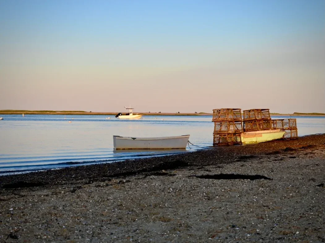 Lobster traps stacked on a boat on a rocky shoreline with boats on calm water and a clear sky in the background.