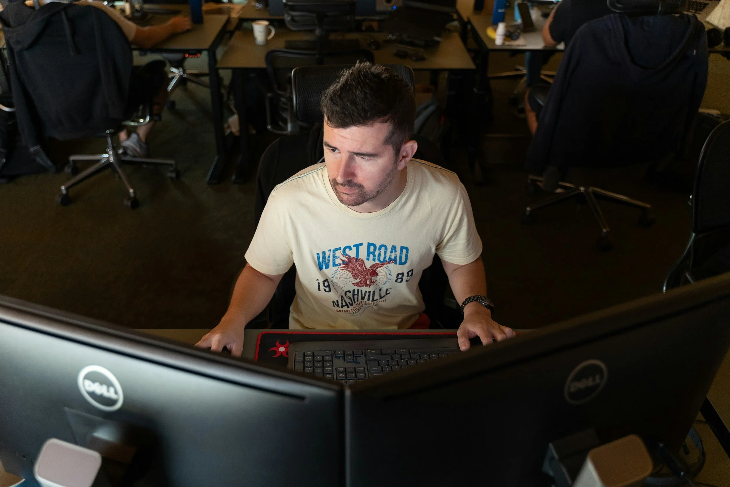 A man sitting at a desk in front of two computer monitors, working on a computer in an office environment with other desks and people in the background.