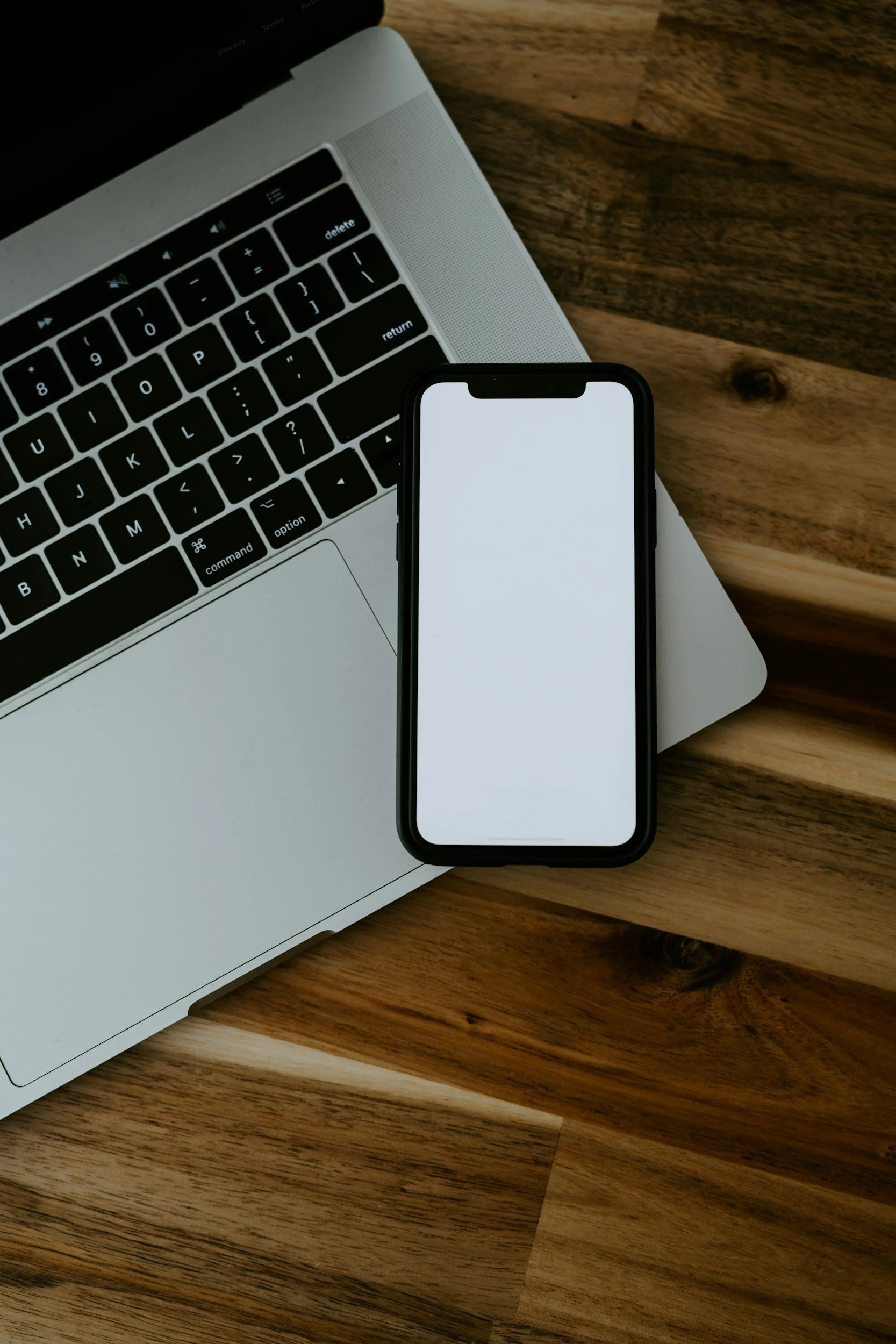 A silver laptop with a black keyboard and a black smartphone with a white screen resting on a wooden surface.