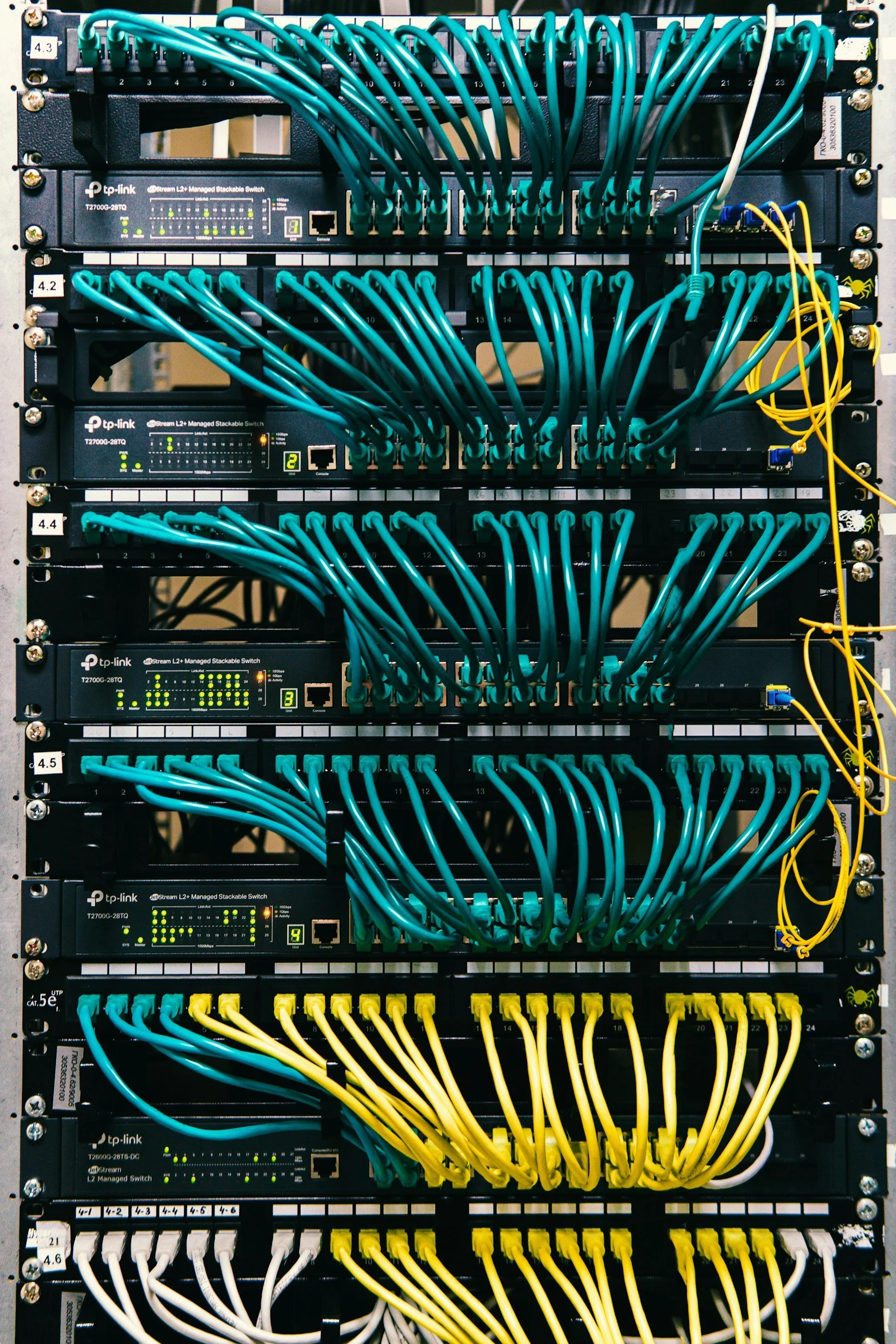 A server rack with multiple network switches, connected by various colored Ethernet cables, including teal, yellow, white, and black.