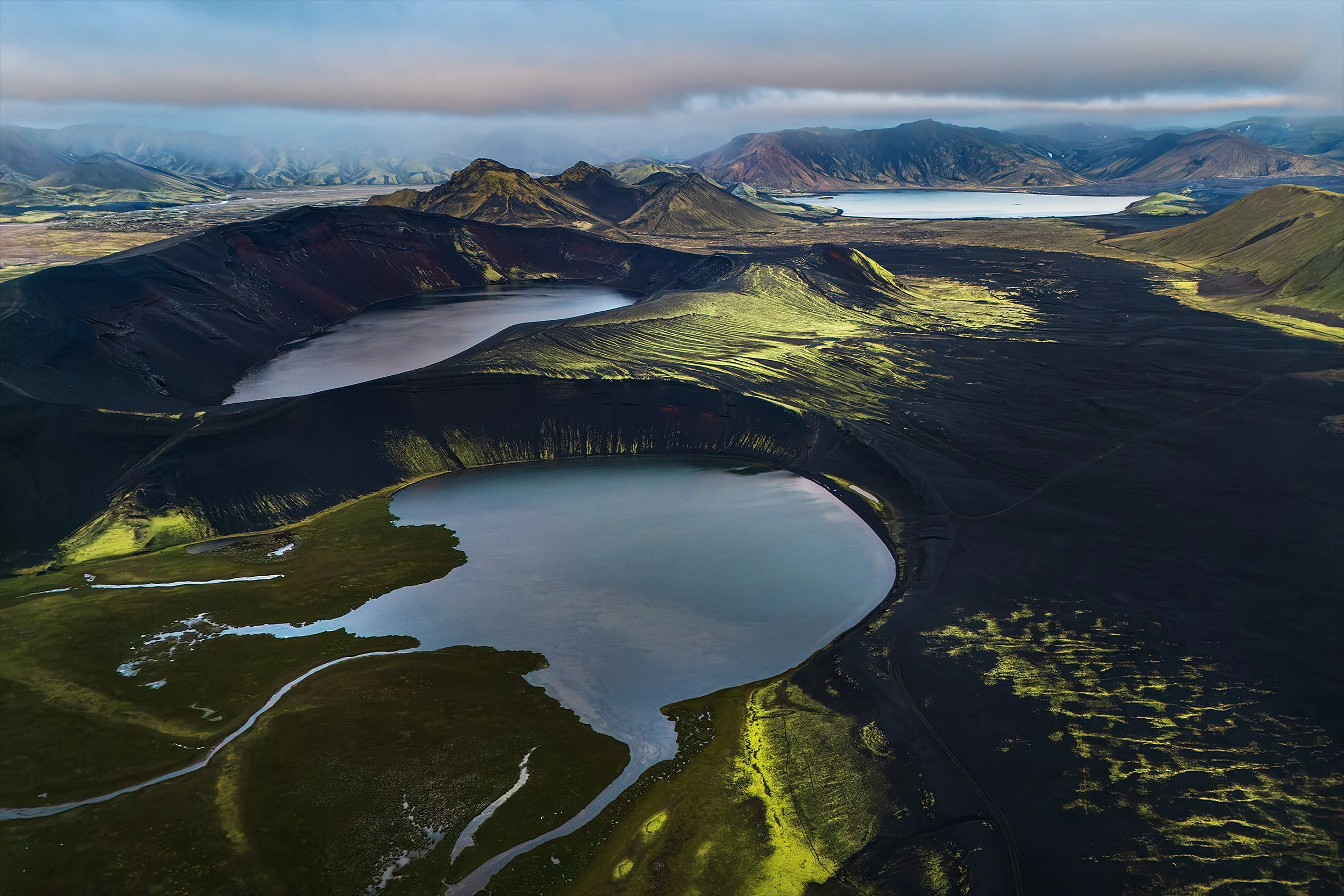 Aerial view of volcanic crater lakes surrounded by green and black volcanic terrain, with distant mountains under cloudy sky.
