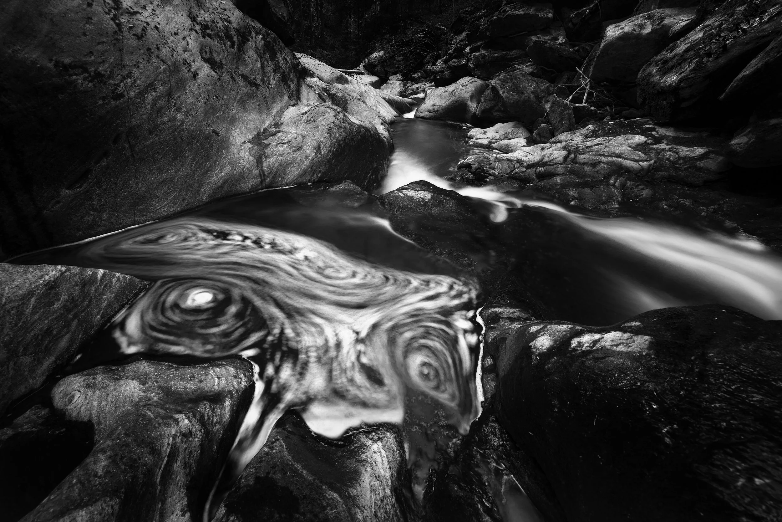 A black and white photograph of a narrow mountain stream flowing over rocks and boulders, with swirling water patterns in the foreground.
