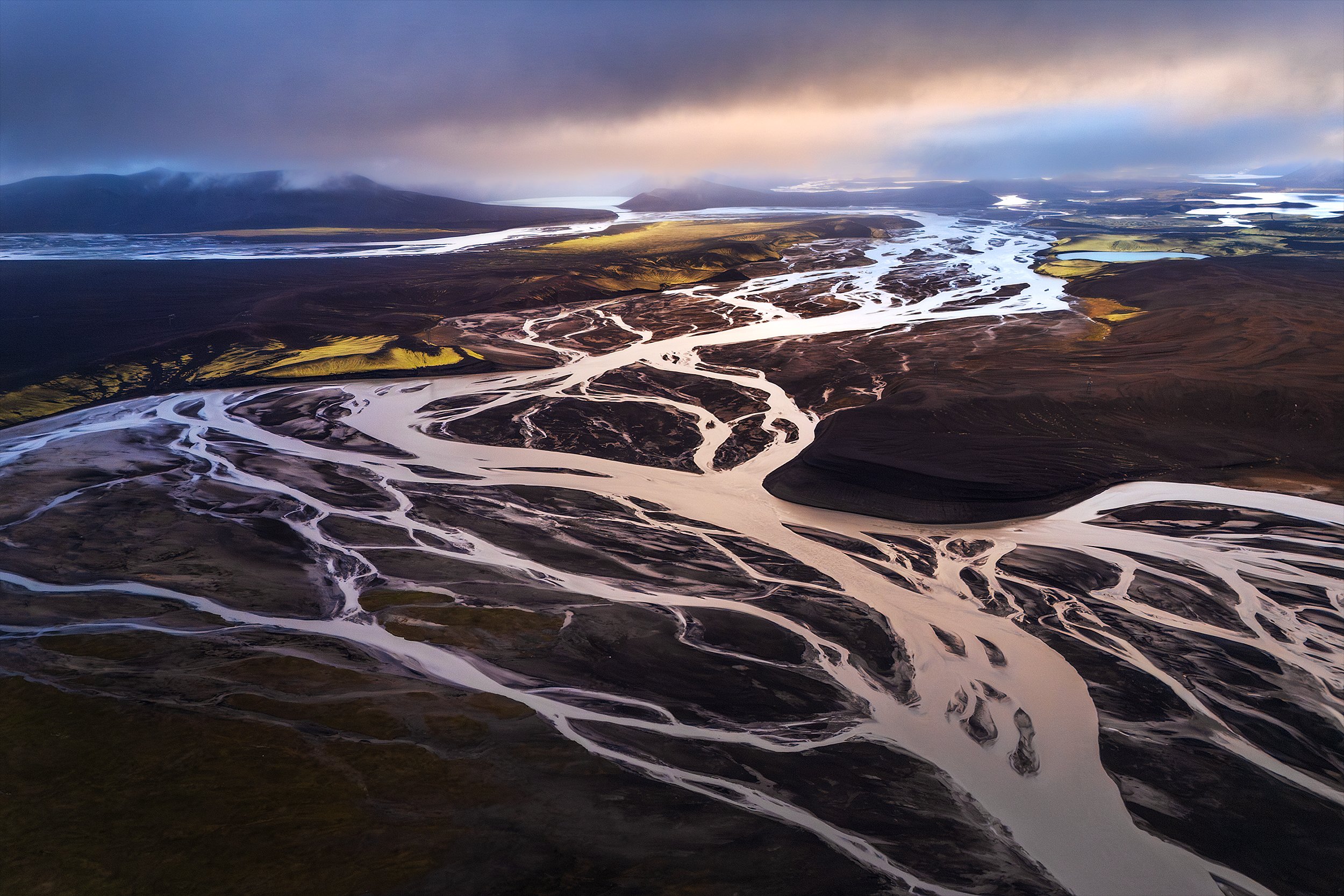 Aerial view of a braided river flowing through a volcanic landscape with mountains in the distance and a cloudy sky.