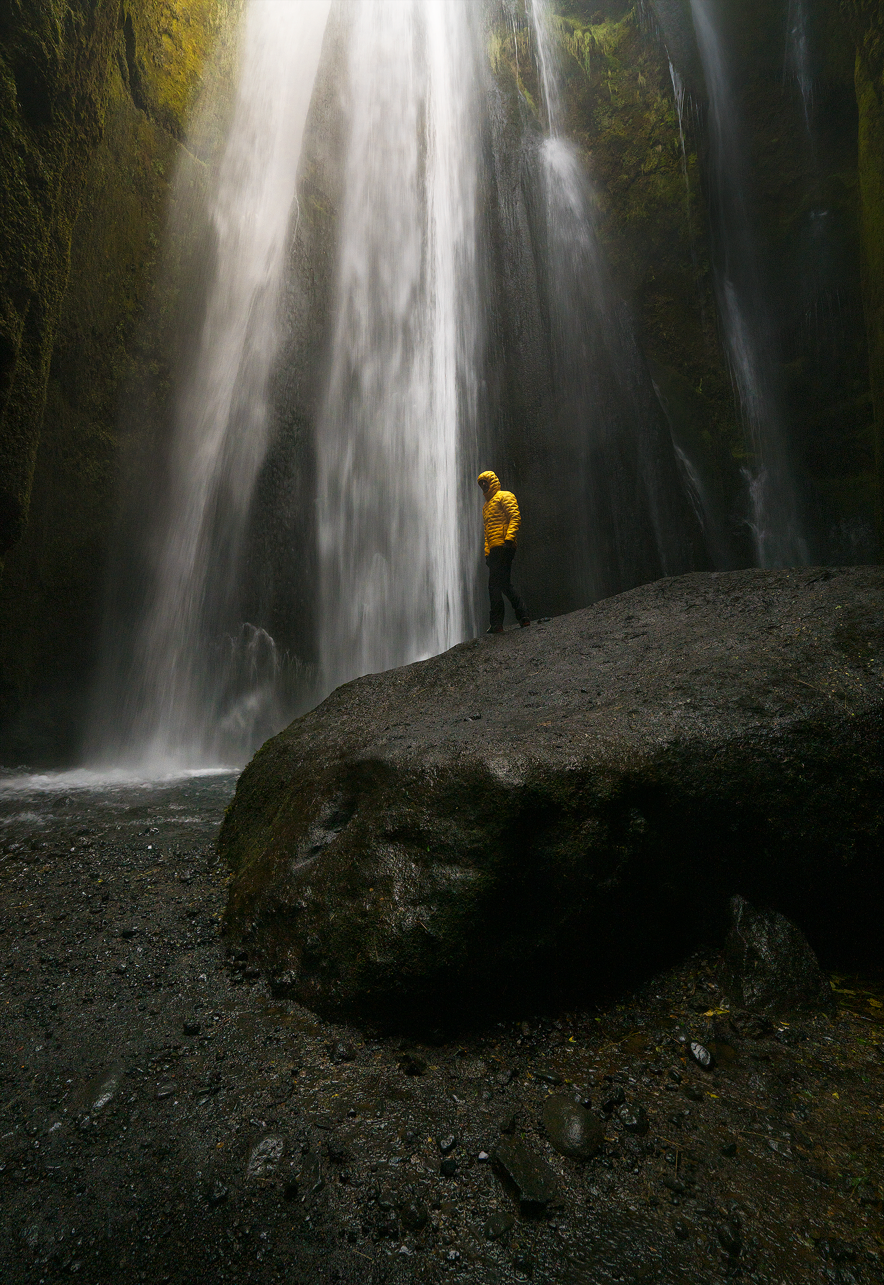 Person in yellow jacket standing on a large rock near a tall waterfall with cascading water and green moss-covered rocks.