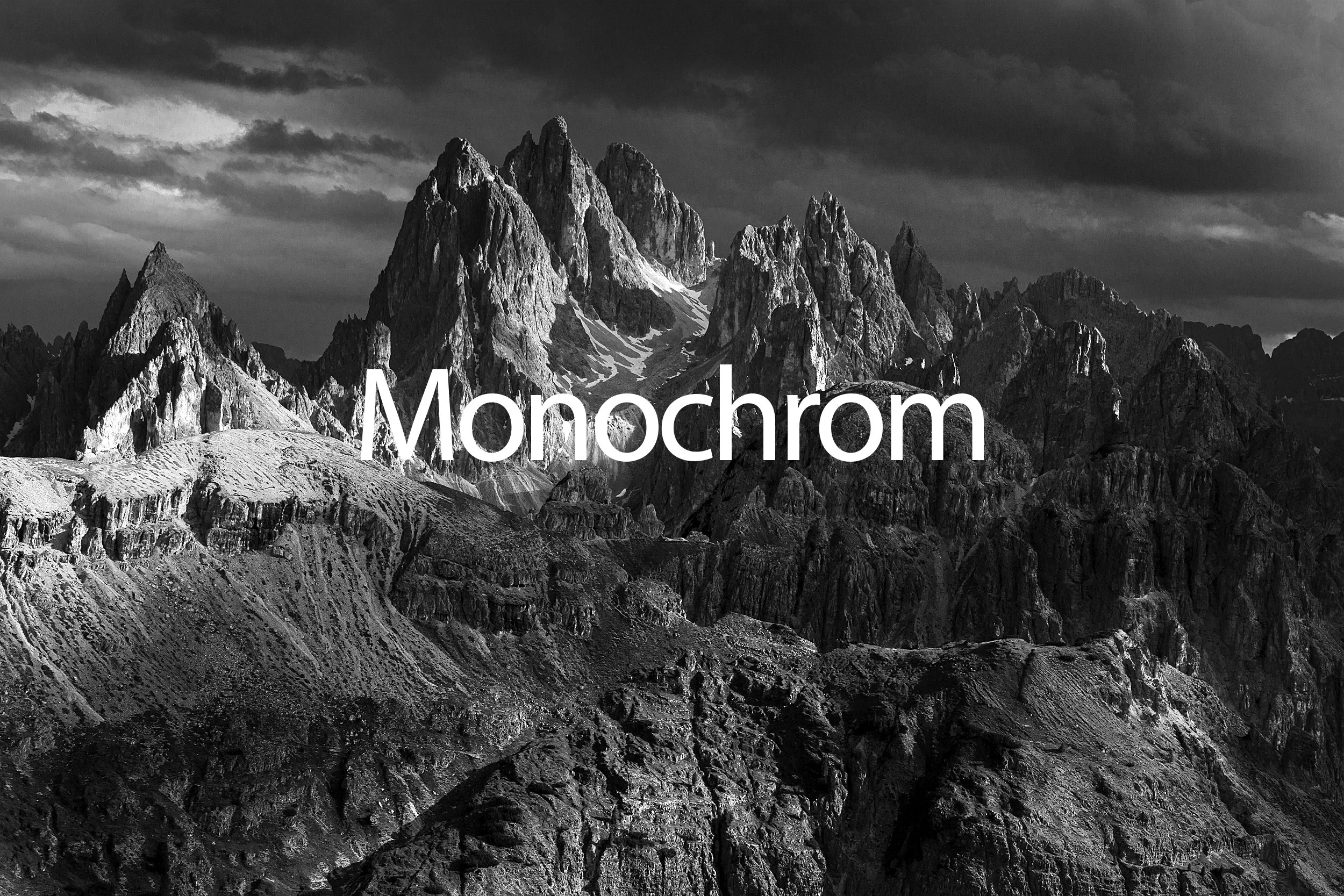 Black and white photograph of jagged mountain peaks with snow patches under a cloudy sky, with the word 'Monochrom' written across the bottom.
