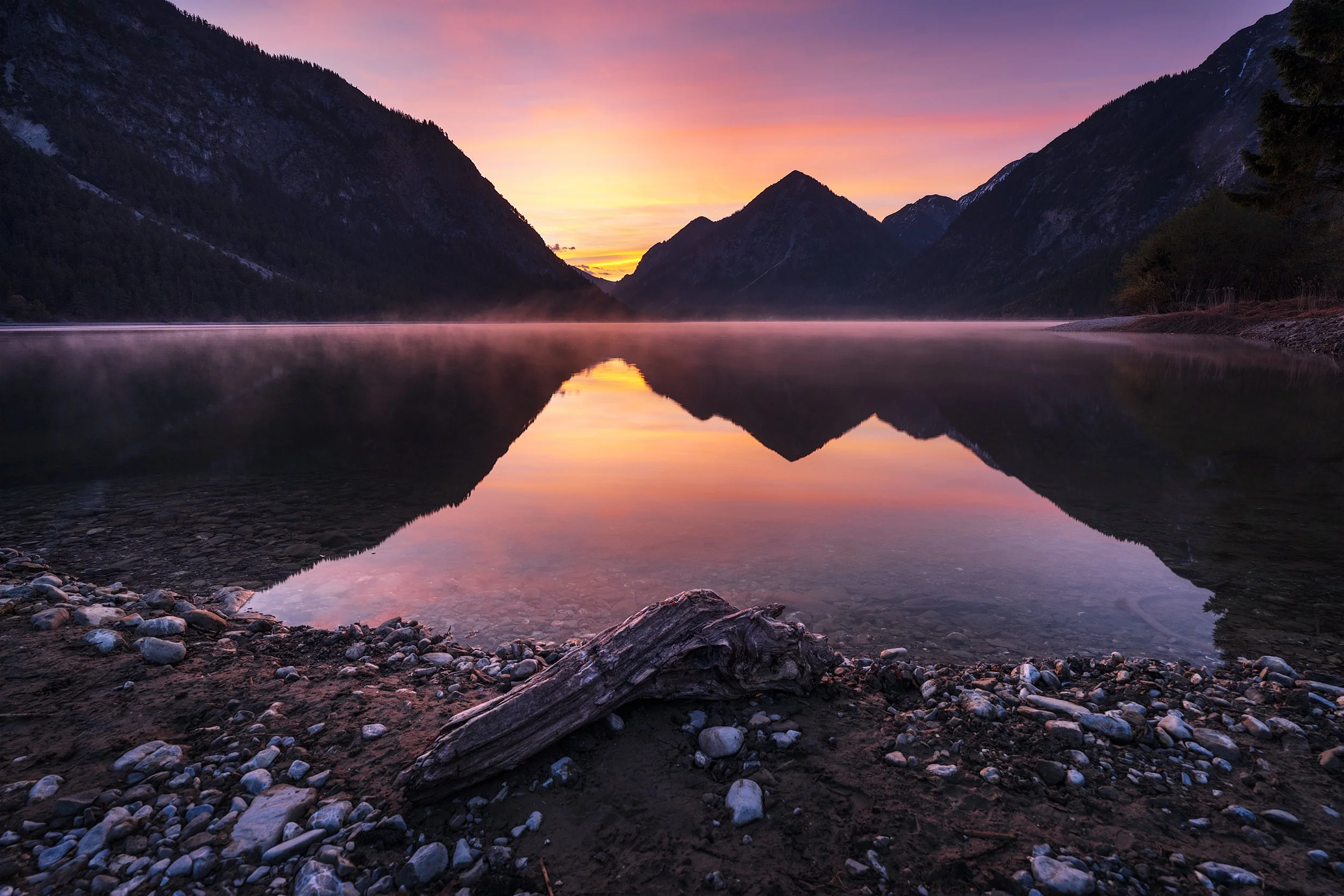 A serene mountain landscape at sunset featuring a calm lake reflecting the colorful sky and surrounding mountains, with rocky shore and some driftwood in the foreground.