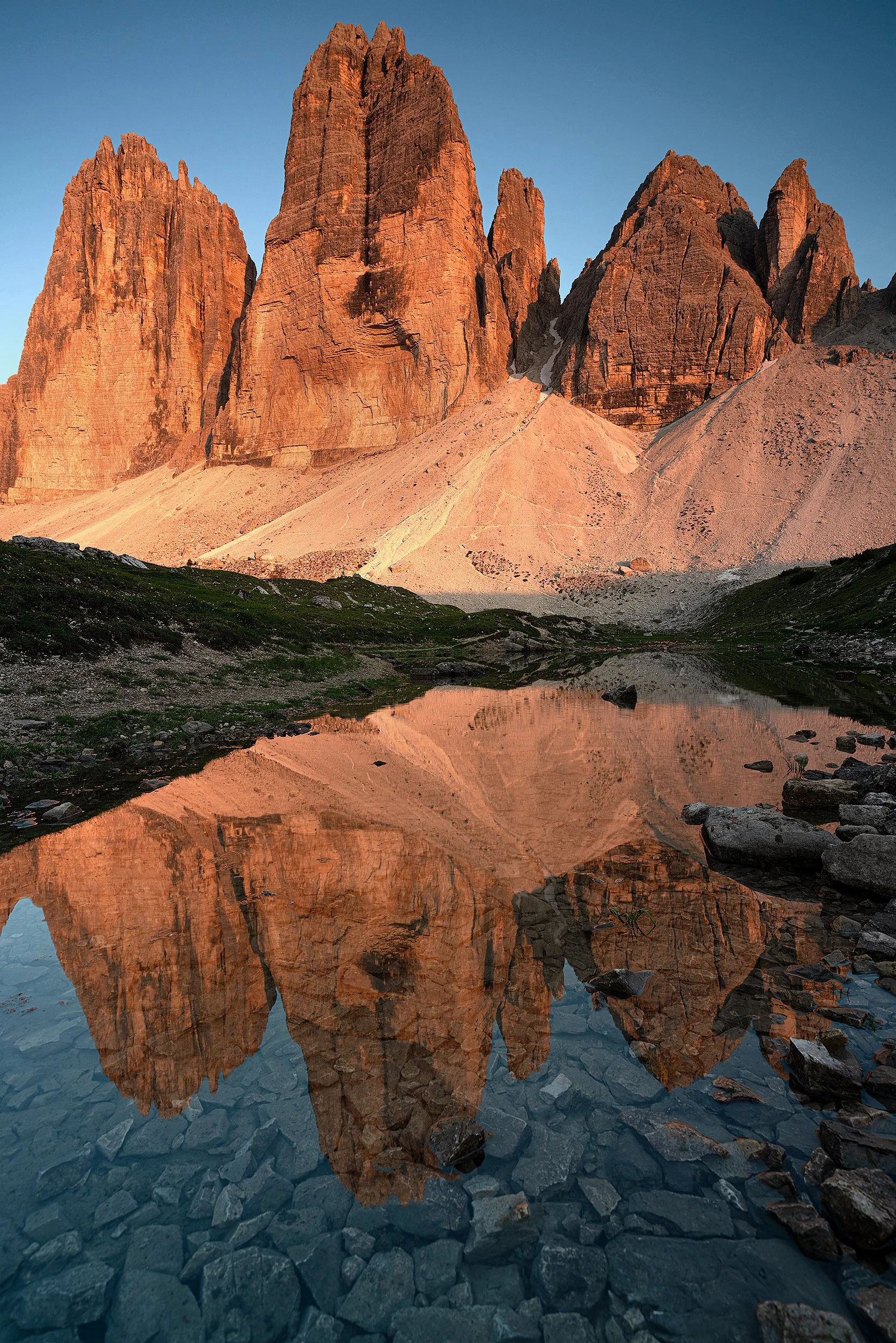 Sunlit mountain peaks reflected in a calm lake with rocky shoreline in a mountainous landscape.