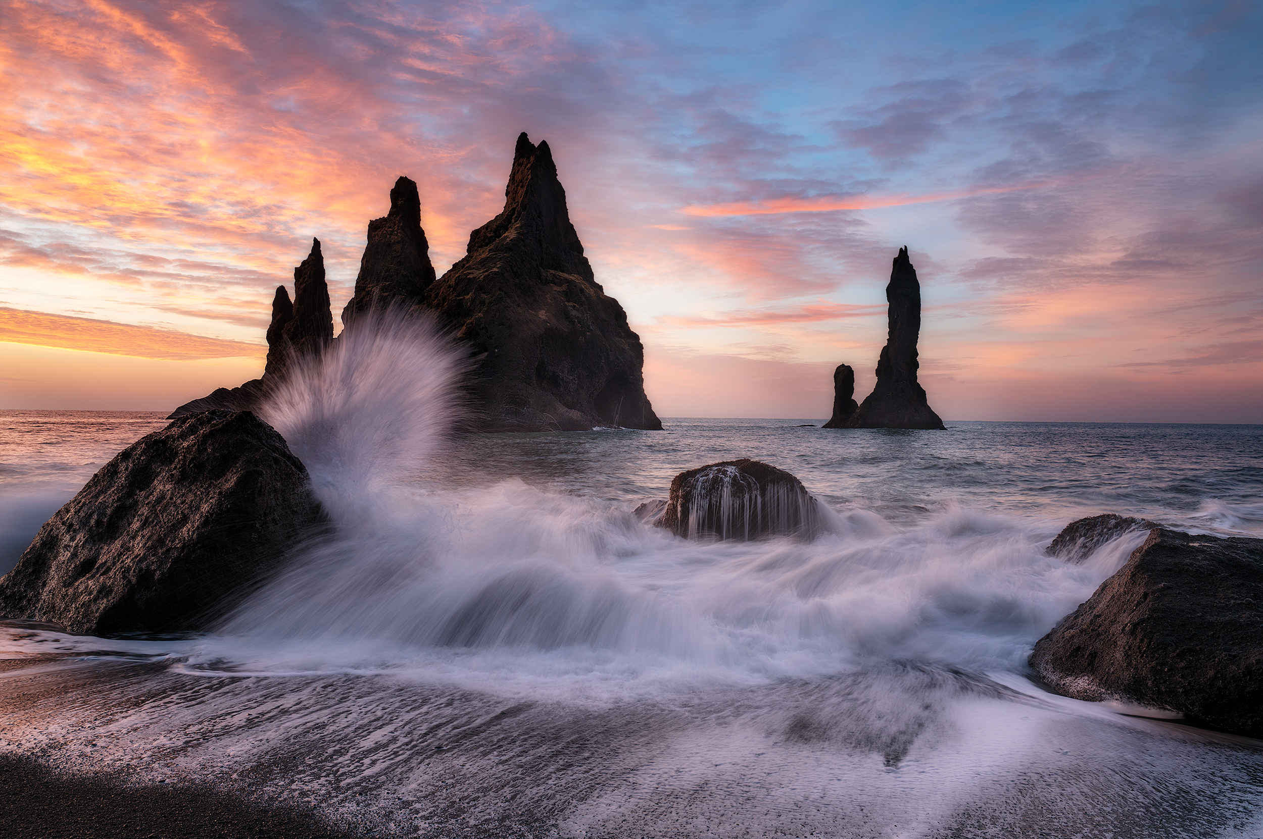 Sunset at the ocean with tall volcanic rock formations, waves crashing on rocks, and colorful sky with clouds